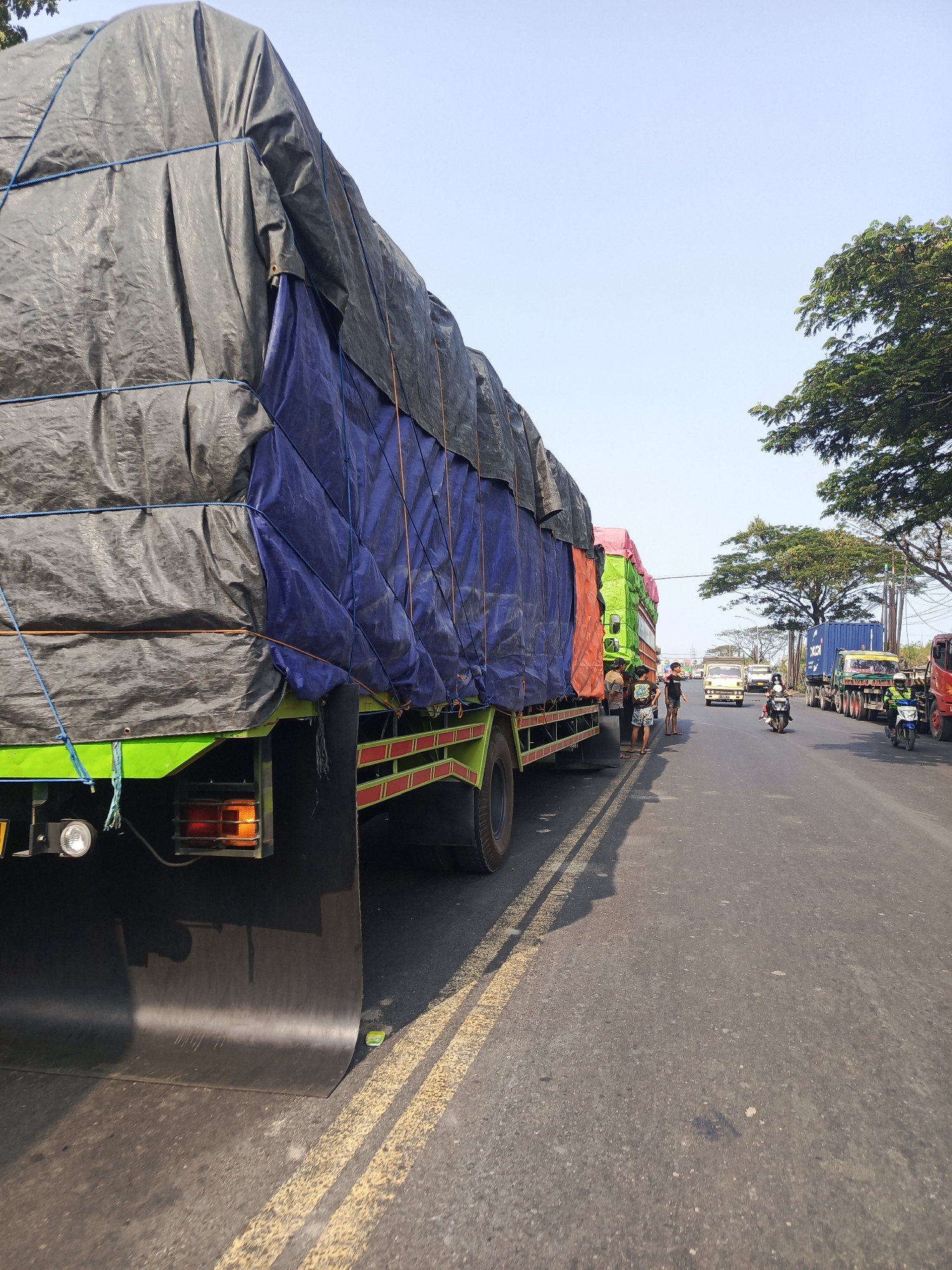 The image captures a traffic scene on a sunny day in Surabaya, Indonesia, likely on a highway or a busy arterial road. The foreground is dominated by the rear of a large green truck, its cargo covered by a black tarp secured with blue ropes. The tarp is draped over a blue and orange tarpaulin, creating a layered effect. The truck's rear lights are visible, with one amber light illuminated. To the right of the truck, the road is asphalt with double yellow lines. In the middle ground, several other trucks are lined up, some also covered with tarpaulins. A few people are standing near the trucks. Further down the road, vehicles including cars, motorcycles, and more trucks, some carrying shipping containers, are moving. Trees line the right side of the road, and the sky is clear blue. The overall impression is one of a busy transportation route with trucks conveying goods.