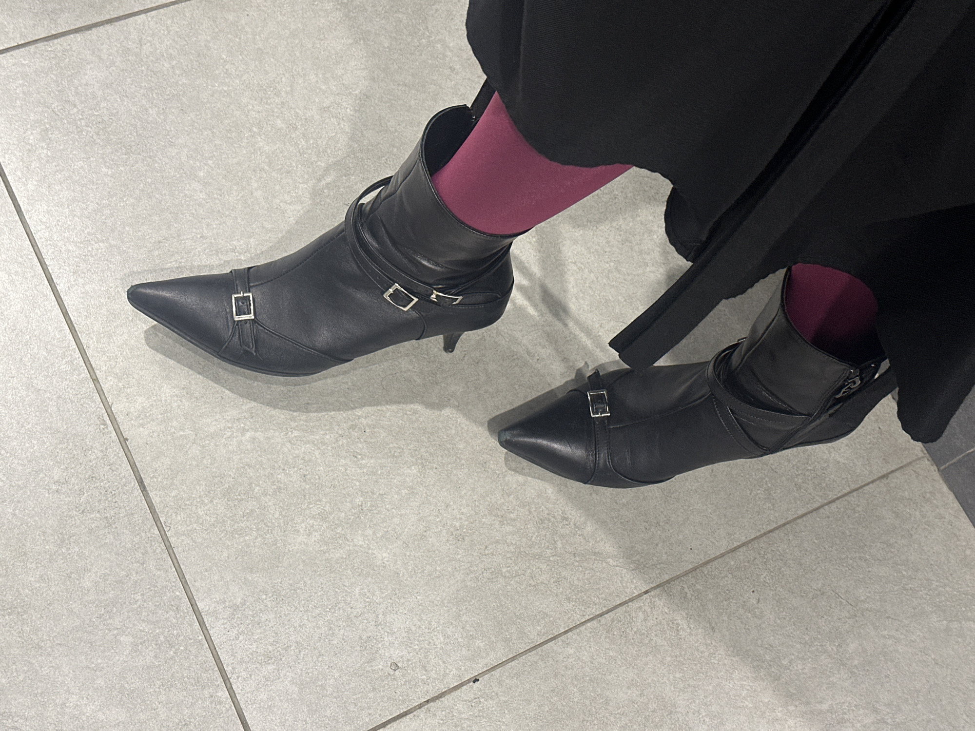 The image shows a close-up of a person's legs wearing black pointed-toe heeled boots with silver buckles and dark pink tights. The person is also wearing a black skirt or dress. The scene appears to be indoors, possibly on a tiled floor. The lighting suggests an indoor environment. There is no visible information to determine the location within Moscow, Russia.