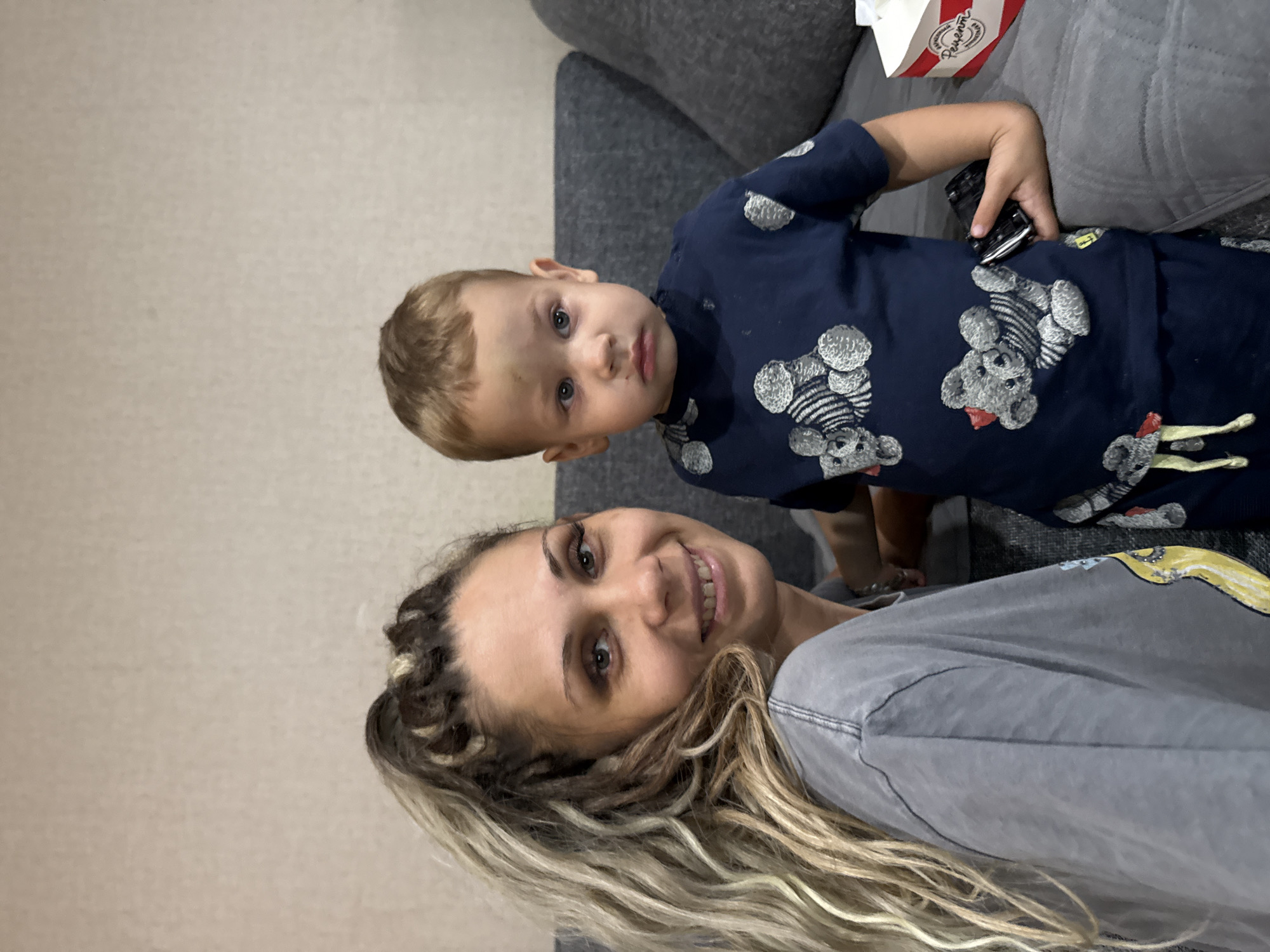 A woman with braided blonde hair and a grey t-shirt smiles at the camera. Beside her, a young boy with light brown hair wearing a dark blue t-shirt with teddy bear prints looks away from the camera. The boy is sitting on a grey sofa. A box of tissues is visible on the right side of the sofa. The background is a plain, light-colored wall. The scene appears to be indoors, likely in a residential setting within Rusanovo, Russia.