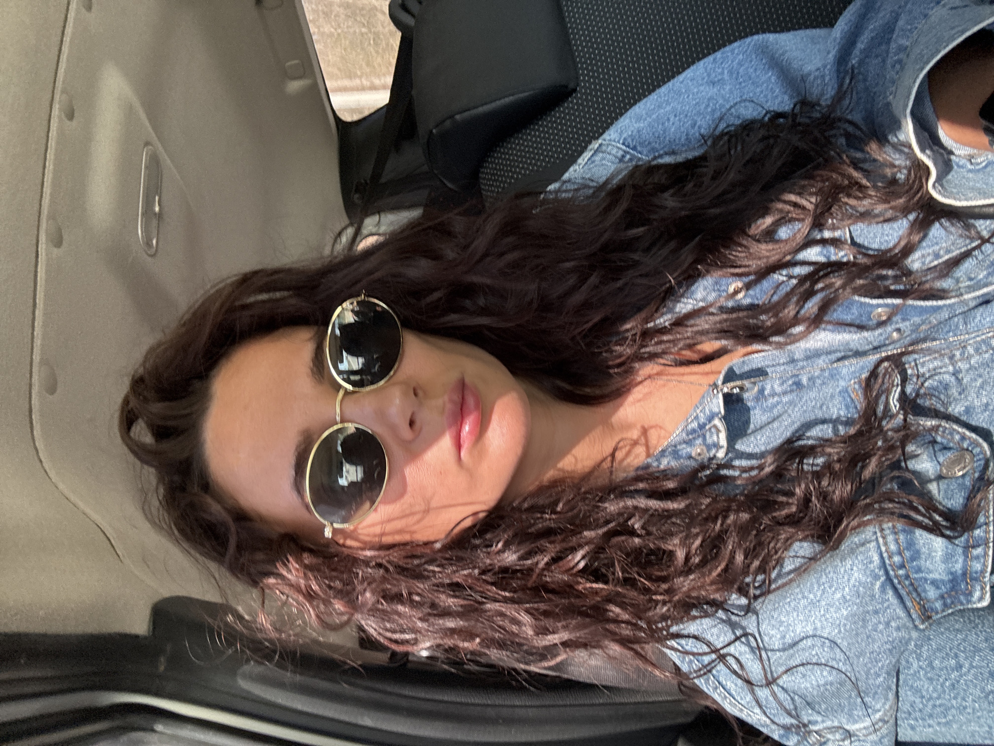 A woman with long, wavy, dark hair is reclining in a car. She is wearing round sunglasses with gold frames and a denim jacket. The car interior includes a grey seat and door panel. There is a black bag behind her. The location cannot be determined to be Chaikovsky, Russia from this image.