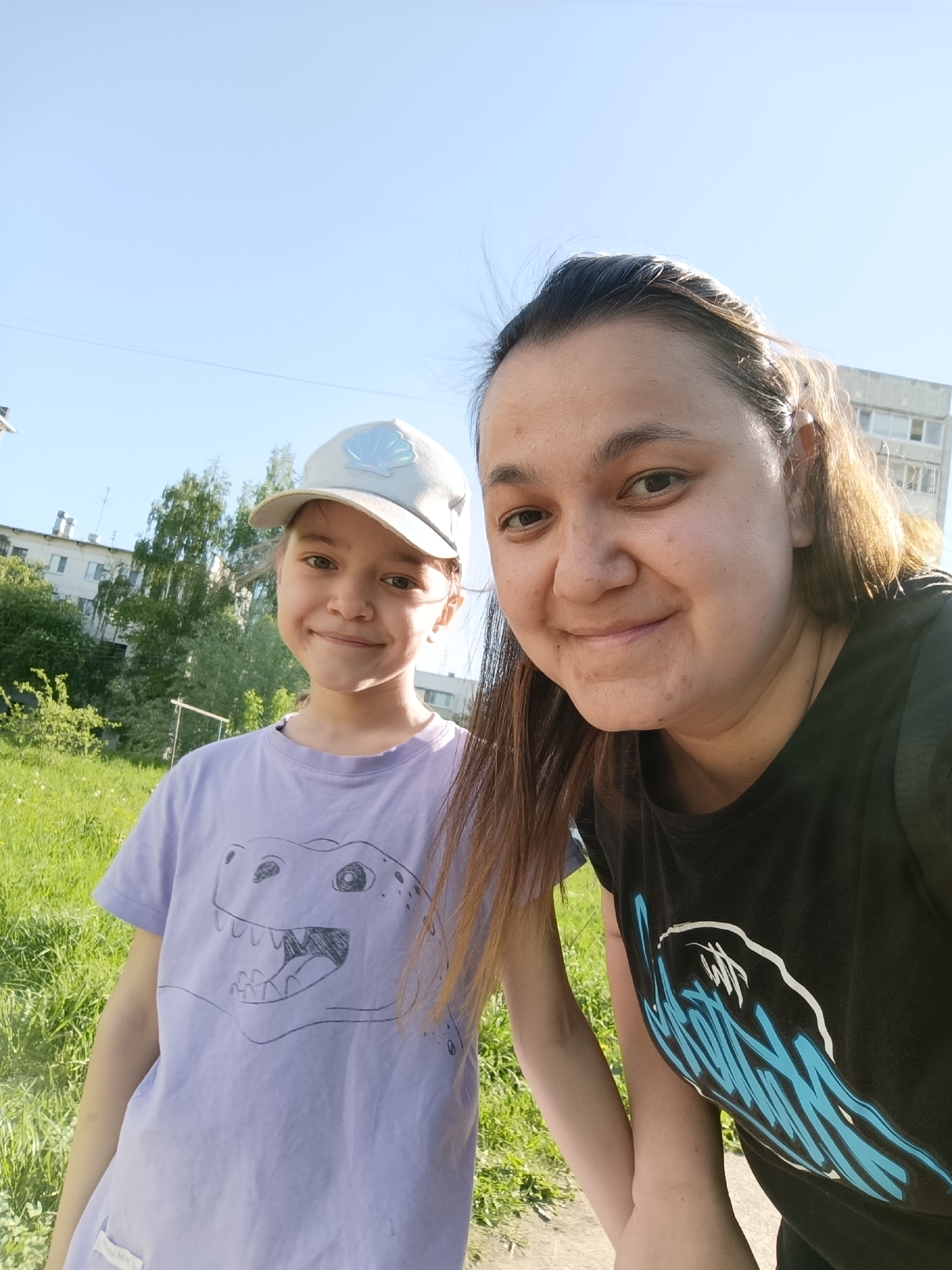 The image is a selfie of a woman and a young girl in an outdoor setting. The woman is on the right side of the frame, with brown hair and a black t-shirt featuring a blue and white design. She is smiling at the camera. To her left is a young girl wearing a lavender t-shirt with a hand-drawn dinosaur on it, and a light-colored baseball cap with a shell design. She is also smiling.
The background consists of a grassy area with some trees and a residential building in the distance. The sky is clear and blue. The location is likely in Chaikovsky, Russia, as indicated by the prompt.
The action is a casual selfie taken by the woman with the girl. They are interacting by posing for the photograph. The environment appears to be a park or residential area.