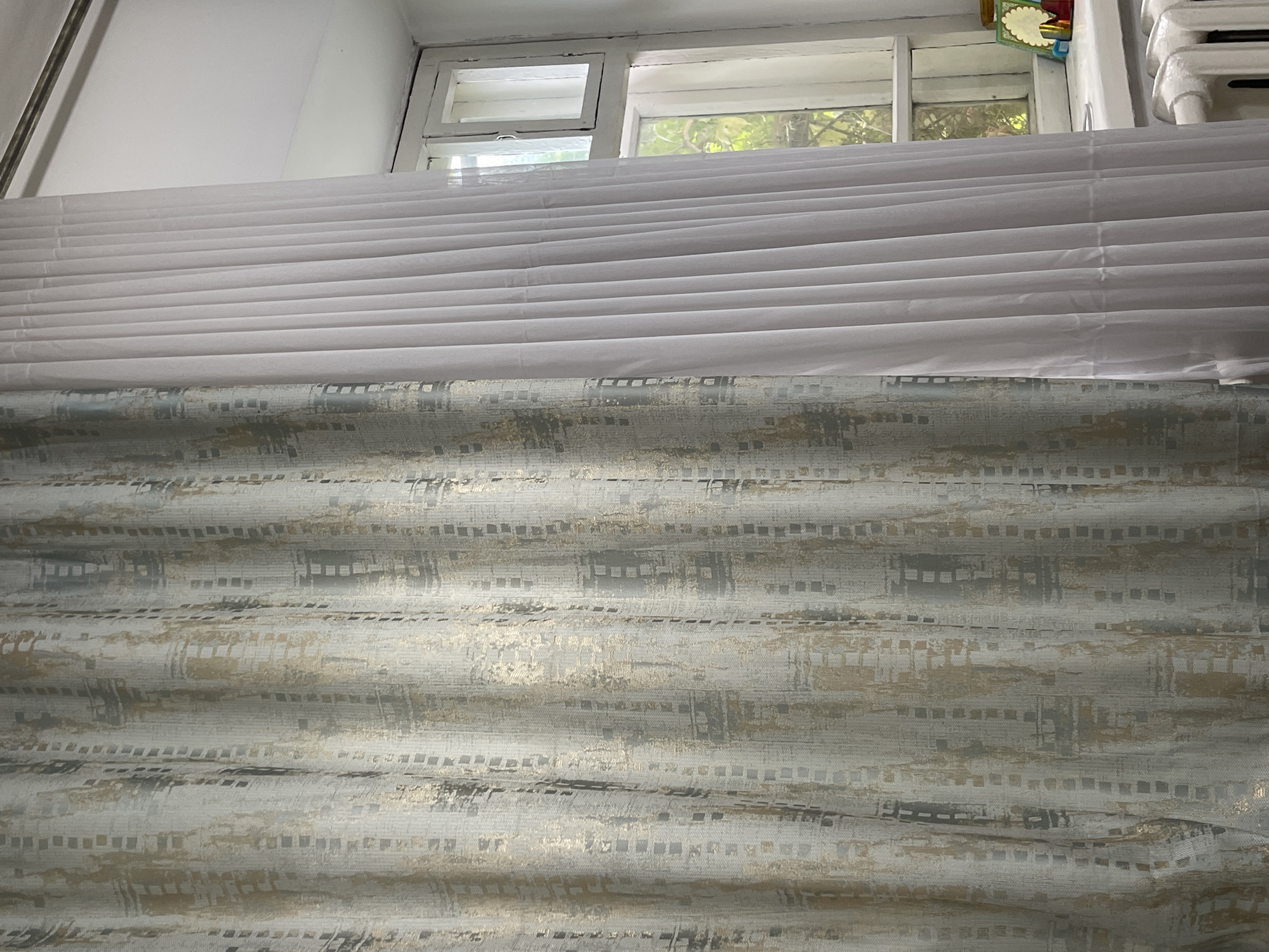 The image shows a window with two layers of curtains. The left curtain has a pattern of gray and gold rectangles. The right curtain is white and sheer. Outside the window, green trees are visible. A white radiator is positioned below the window. A small, framed picture and other objects are placed on the windowsill. The scene appears to be an indoor setting, likely a room in a building. The location is not specified as Bishkek City, Kyrgyzstan.