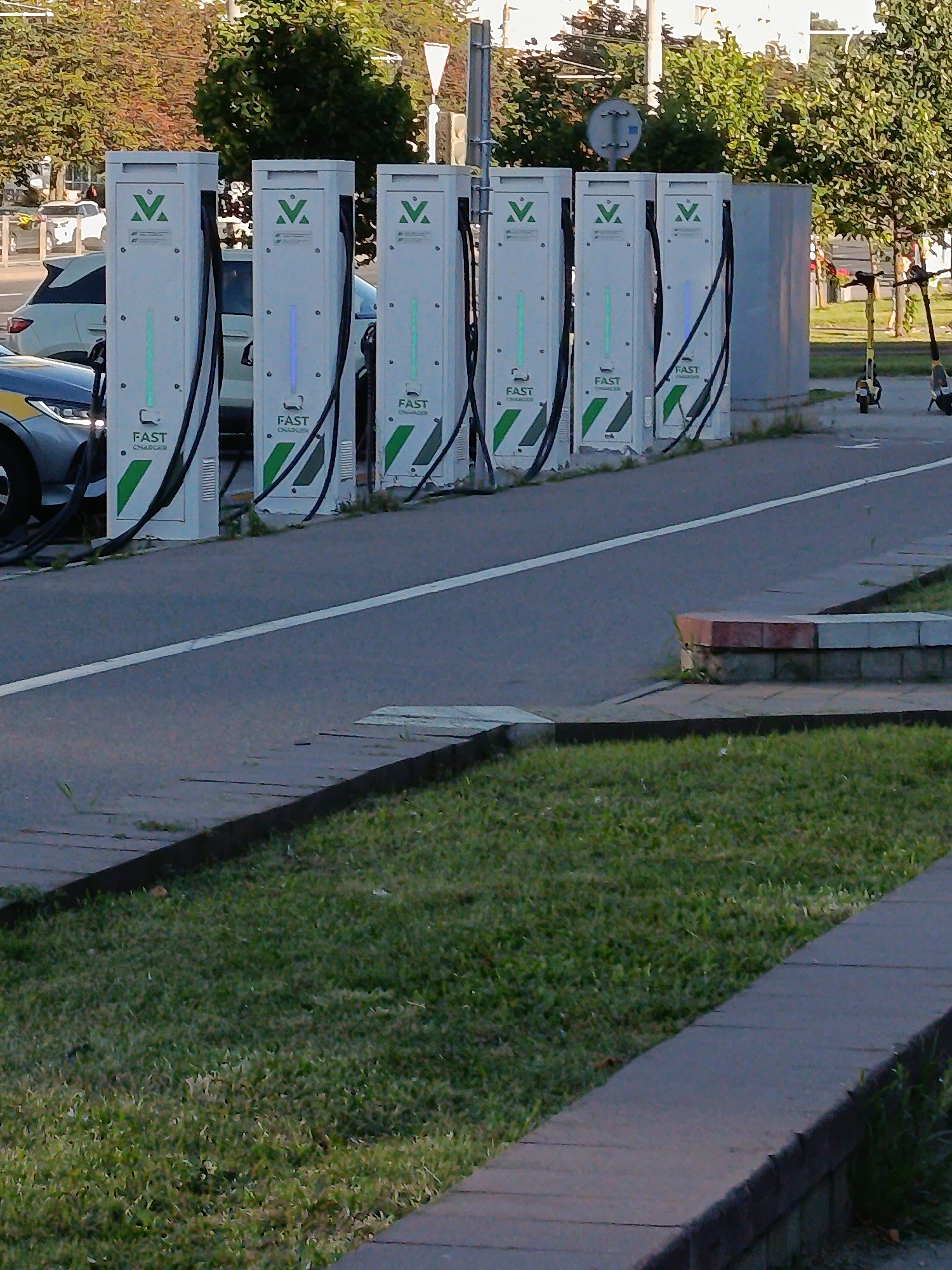 The image shows a row of five electric vehicle charging stations lined up along a road. The charging stations are tall, white, and rectangular, with a green and white "V" logo at the top. Below the logo, each station has a display that reads "FAST CHARGER". Some of the stations have illuminated green or blue lights. Cables hang from each station, ready to be plugged into vehicles.
In the foreground, a portion of a car is visible, parked next to one of the charging stations. The car appears to be dark-colored with a yellow stripe on its side. Further in the background, two electric scooters are parked, one yellow and one black. Behind the charging stations and scooters, there are trees with green foliage, suggesting a pleasant, possibly spring or summer day. A traffic signal and a white triangular sign are also visible in the background. The scene is set on a paved area adjacent to a road with a white dividing line. There is a grassy area with a stone border in the foreground.
The image captures a modern urban setting, likely a charging point for electric vehicles and scooters, indicating a focus on sustainable transportation. The lighting suggests it is daytime, with the sun casting shadows. No people are visible in the image, making the focus solely on the objects and the setting. The text "FAST CHARGER" is clearly legible on all the charging stations. The location context provided is Minsk, Belarus.