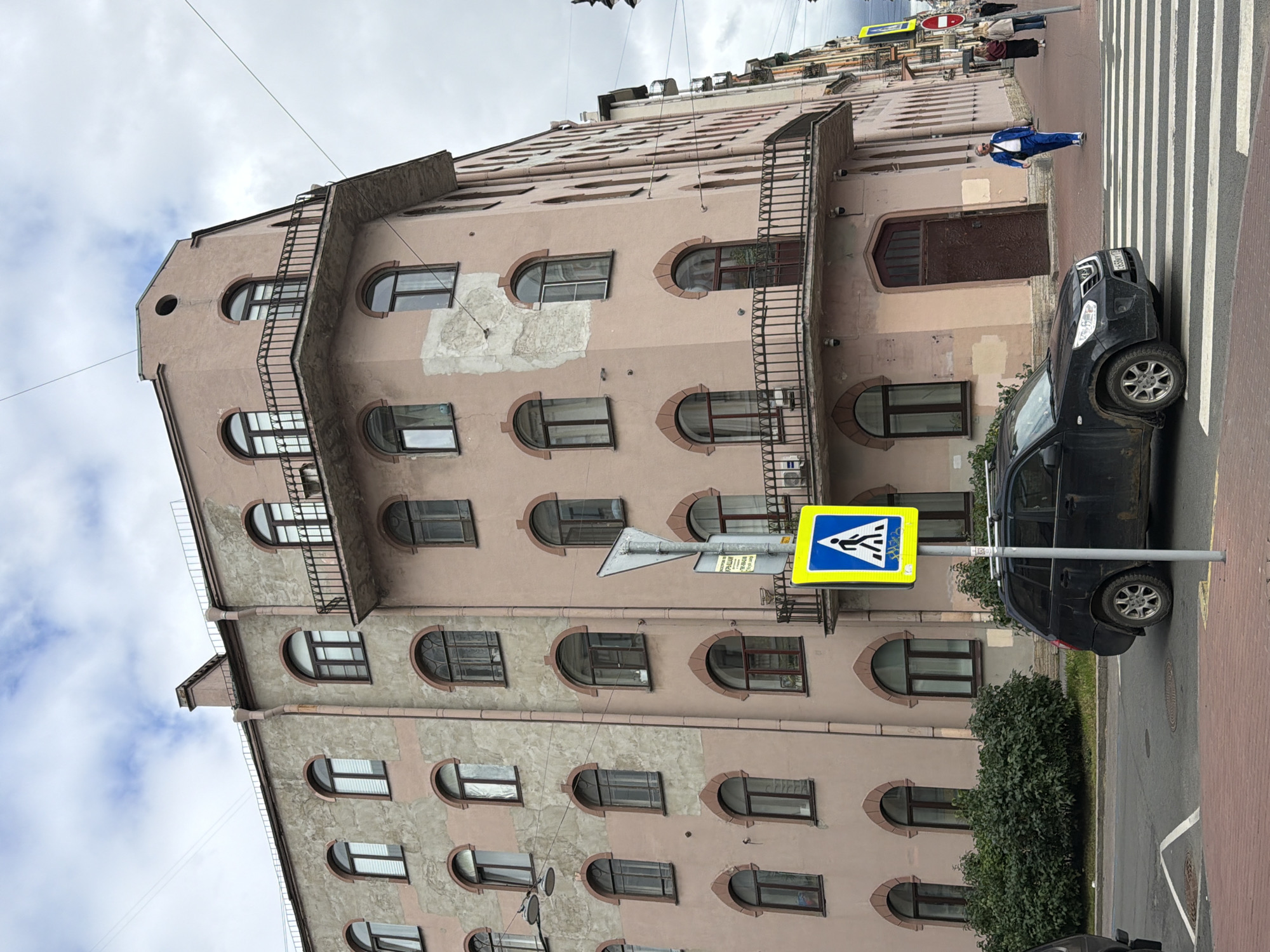 The image captures a street scene in Saint Petersburg, Russia, under an overcast sky. A multi-story building with a pale pink facade, featuring arched windows and decorative detailing, dominates the frame. The architecture suggests an older style, possibly Art Nouveau or a related historical period. A black hatchback car is stopped at a pedestrian crossing, with its front partially obscuring the crossing lines. A pedestrian crossing sign, a yellow square with a blue pictogram of a person walking on a crosswalk, is positioned on the right side of the road.
Further down the street, several people are walking on the sidewalk. One man, dressed in a bright blue tracksuit and white sneakers, walks towards the right of the frame. Two women, one in a dark dress and the other in lighter clothing, are also visible on the sidewalk. Another red and white circular sign is visible further down the street. Green bushes line the base of the building in the foreground. The street itself is paved with red brick on the sidewalk and asphalt on the road. The overall atmosphere is that of a typical urban environment on a cloudy day.