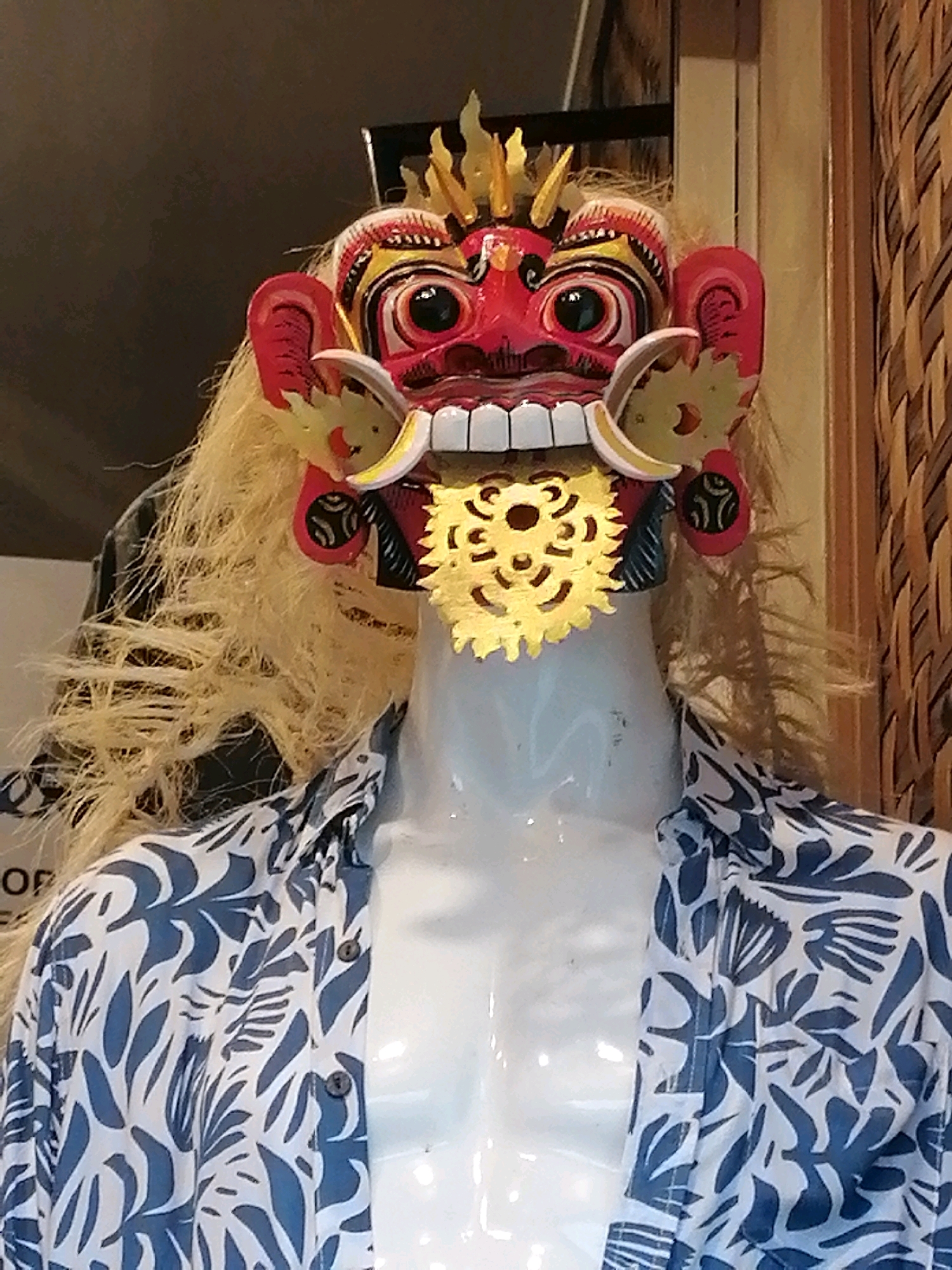 This is a medium shot of a traditional Balinese mask displayed on a white mannequin. The mask is intricately designed with red, yellow, and black colors. It features large, wide eyes, a gaping mouth with white teeth and a gold circular ornament in the center, and flame-like yellow protrusions on the forehead and ears. Long, blonde hair extends from the mask, framing a white mannequin neck and chest adorned with a white shirt featuring a blue floral pattern. The background consists of a dark, textured surface, possibly a wall or screen, and a woven bamboo pattern. The setting appears to be a shop or display area in Ubud District, Indonesia, given the cultural context of the mask.