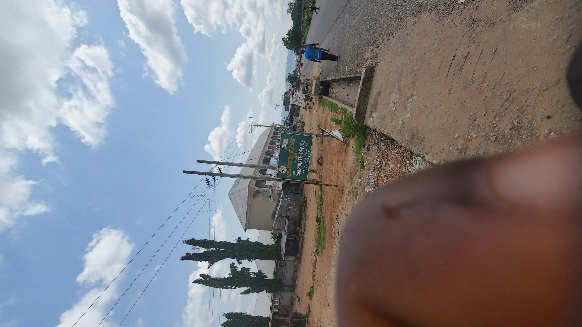 In the foreground, a blurred finger partially obstructs the view. The scene depicts a street in a developing area, with a paved road running along the right side of the image. On the left side of the road, there is a dirt area with scattered debris, including what appears to be discarded tires and small structures. Further back, a multi-story building with a light-colored facade and a covered walkway is visible. A green signpost stands in front of the building, indicating "NIMET" and "NIGERIAN METEOROLOGICAL AGENCY (NIMET) CORPORATE OFFICE". Several tall, thin trees line the left side of the scene. Power lines are strung across the sky, supported by utility poles. A few people are visible walking along the road, with one person in a blue shirt walking away from the camera. The sky is partly cloudy with scattered white clouds against a blue backdrop. The overall impression is of an urban or semi-urban environment in a region with infrastructure that is still under development.
