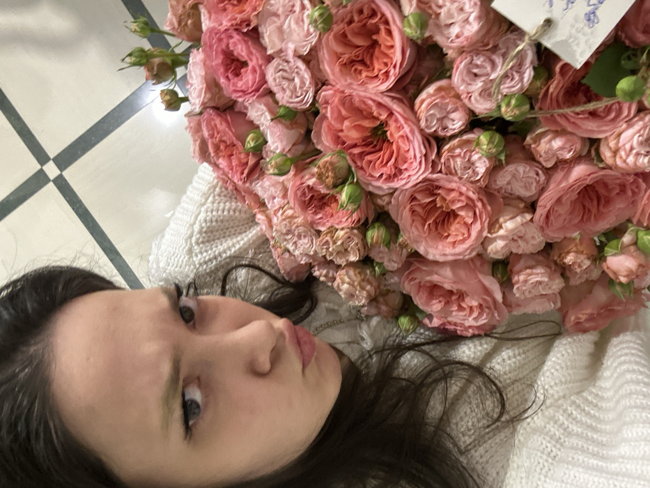 A woman lies on a white sweater, looking to the side with a slight pout. Her dark hair surrounds her face, contrasting with her fair skin. A large bouquet of pink roses partially covers her, with some buds visible. The background includes a tiled floor with a gray and white pattern.