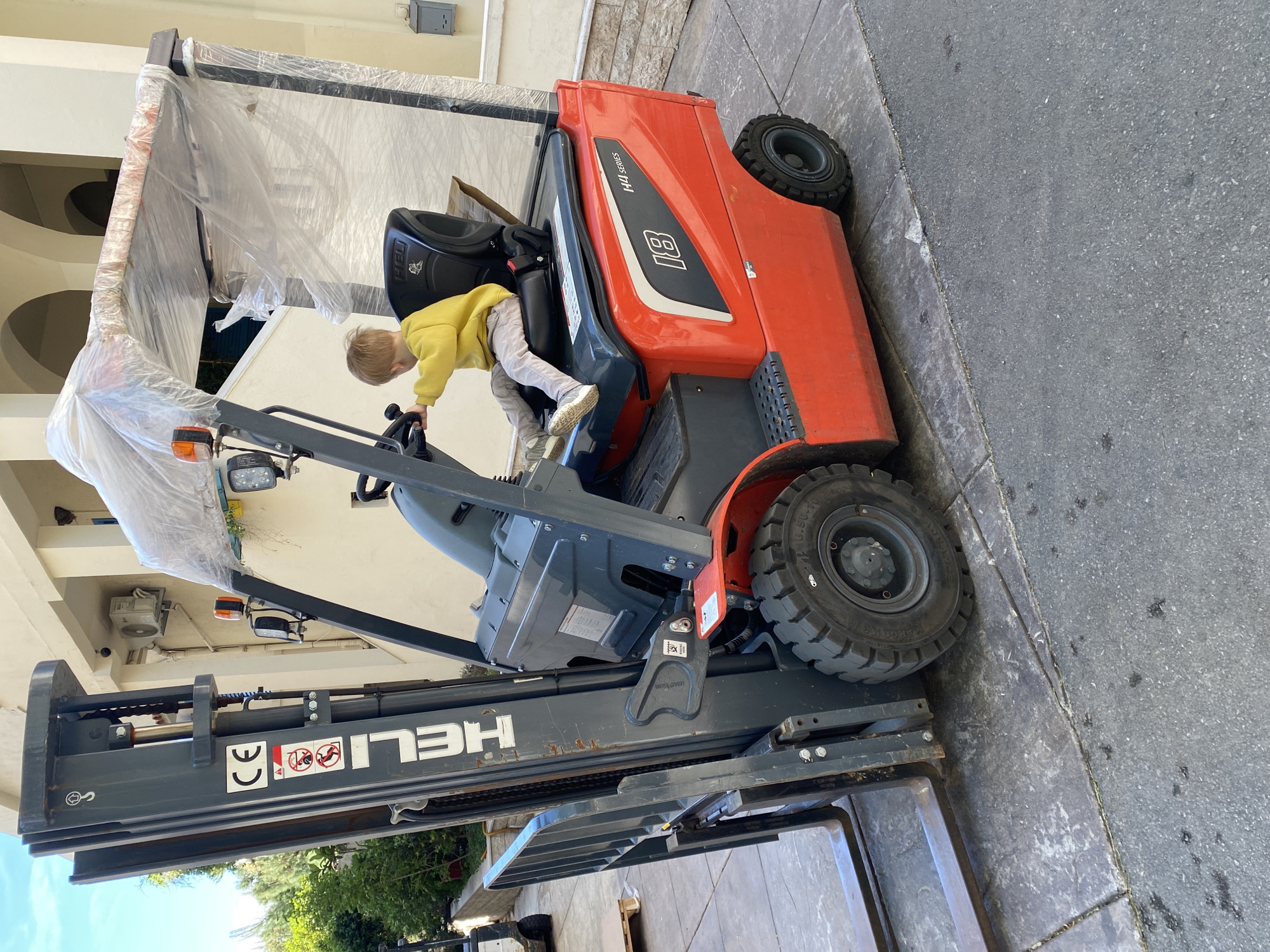 A child in a yellow shirt and light-colored pants is climbing onto a red and gray forklift. The forklift is parked on a paved surface next to a building. A covered object is attached to the top of the forklift. The "HELI" logo is visible on the gray part of the forklift. The surrounding environment includes greenery and what appears to be a building exterior. The location is Rafailovići, Montenegro.