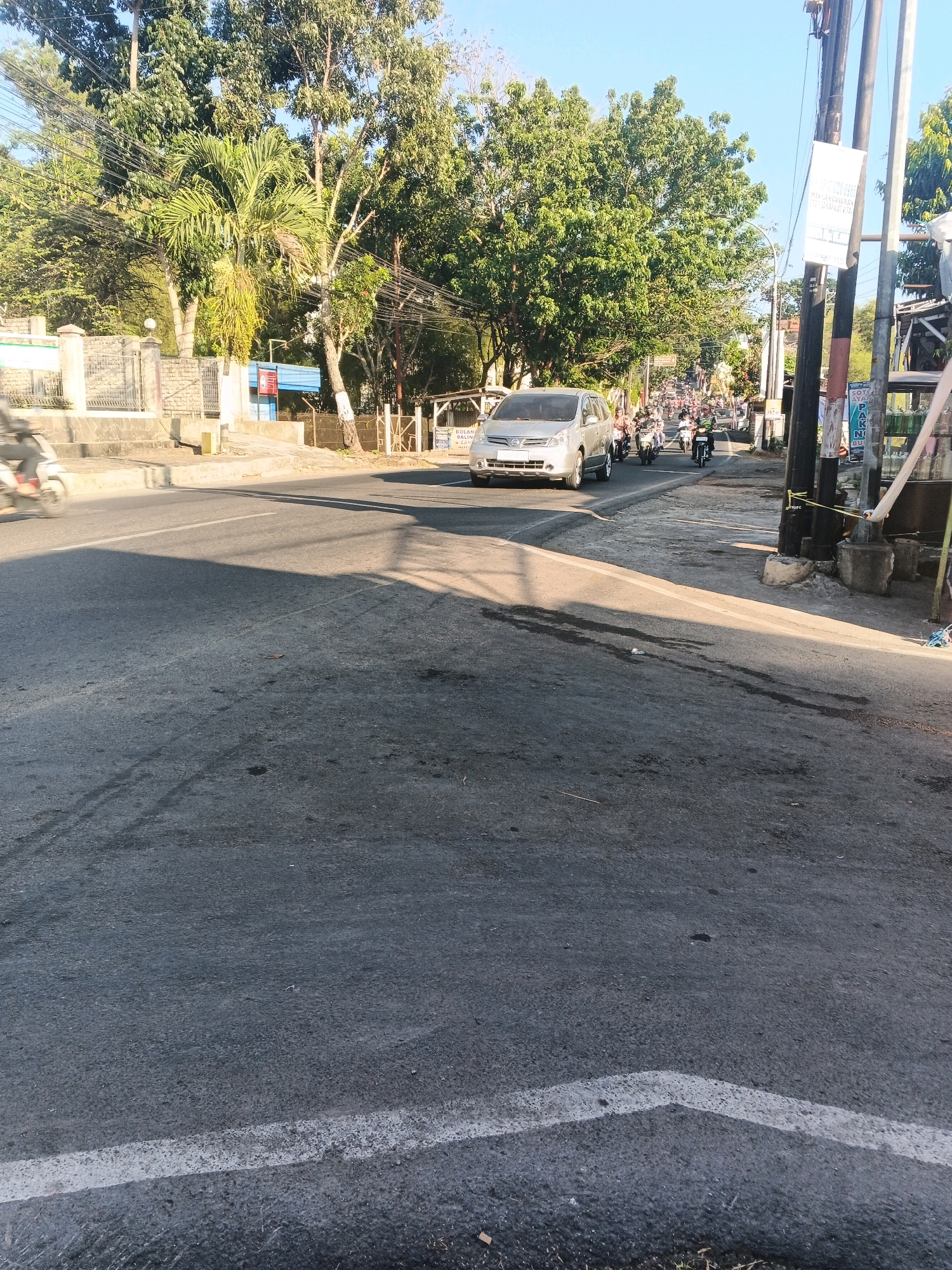 The image depicts a street scene in Semarang, Indonesia, likely during daylight hours. A silver minivan is visible in the foreground, traveling on the right side of the road. Several motorcycles are also present, forming a traffic flow further down the street. The road is paved and appears to be a moderately busy thoroughfare, with trees and buildings lining the sides. Power lines are strung overhead, supported by utility poles. The sky is clear and blue. In the immediate foreground, the asphalt road surface is visible, showing some wear and tear, including dark patches and a white lane marker. A motorcycle is seen in motion on the left side of the frame, blurred due to its movement.
