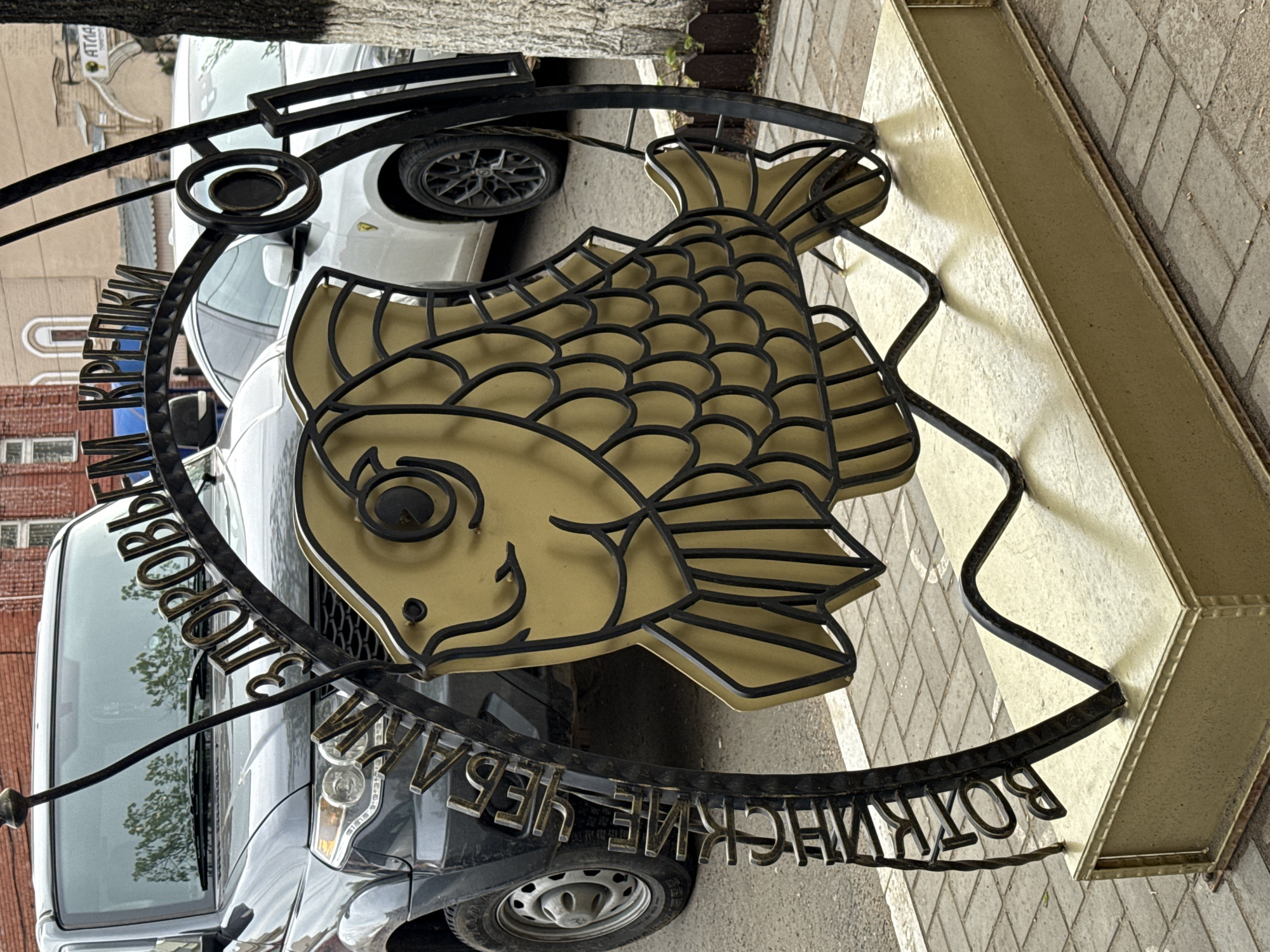 An eye-level outdoor shot shows a metal sign with a cartoon fish in Votkinsk, Russia. The sign is gold with black outlines and a black frame. The fish has a smiling expression. Russian Cyrillic script is visible around the perimeter of the circular frame. A gray car and a white car are behind the sign. Brick buildings and a tree trunk are in the background. The sign is on a narrow paved street.
