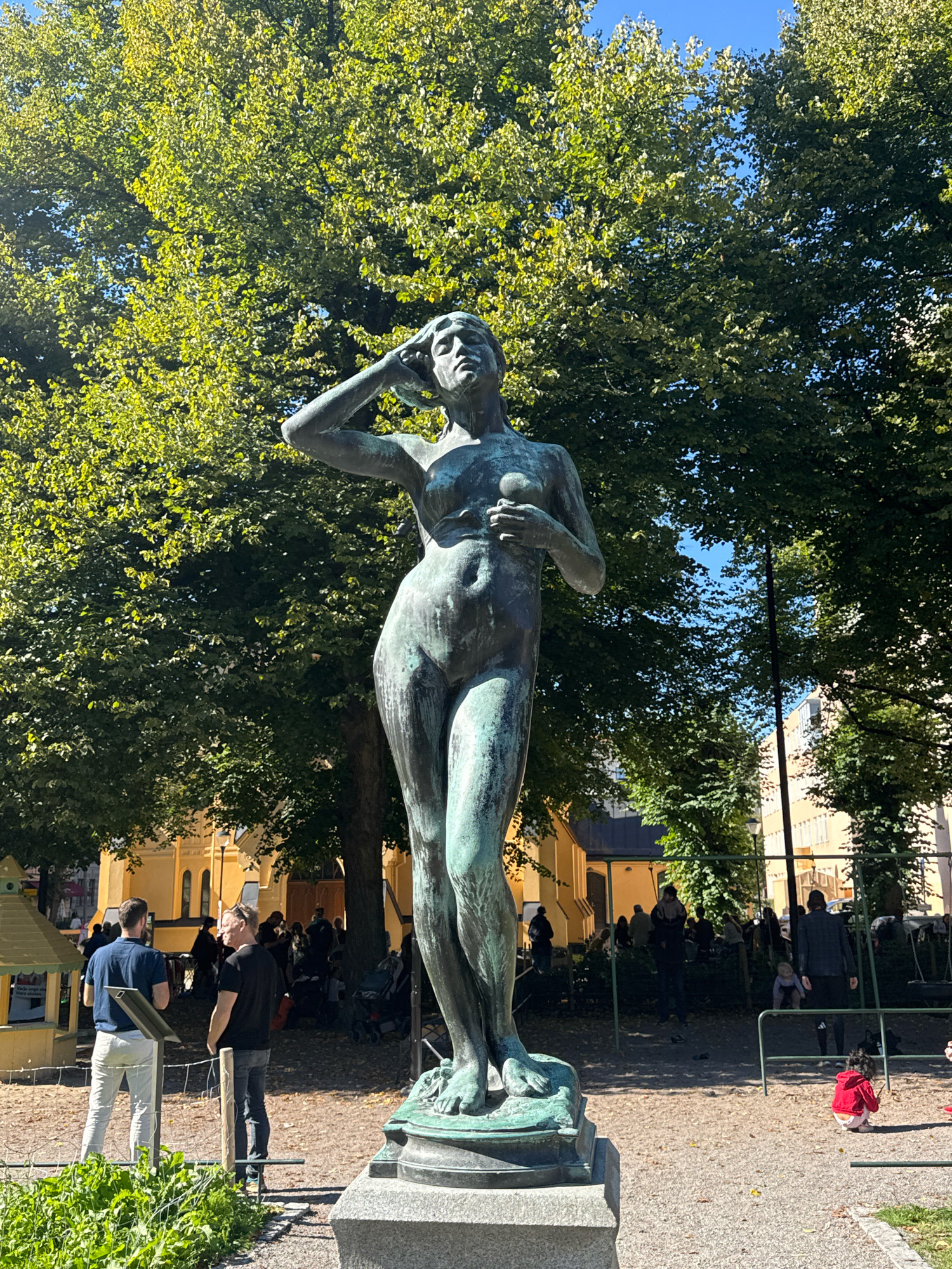 The image depicts a bronze statue of a nude woman in a park setting in Stockholm, Sweden. The statue is positioned on a stone pedestal, with its right arm raised to its head and its left hand holding a small sphere. The statue's patina suggests it is aged, with green oxidation visible on its surface.
Behind the statue, lush green trees with dense foliage dominate the background, with patches of blue sky visible through the leaves. Sunlight filters through the trees, casting dappled shadows on the ground.
In the middle ground, several people are scattered throughout the park. On the left, two men are standing near a yellow playhouse structure, one in a blue shirt and white pants, the other in a black t-shirt and jeans. Further back, a yellow building with arched windows is visible, possibly a church or public building. A playground with swings and a slide can be seen on the right side of the image, with several children and adults present. One child in a red jacket is sitting on the ground near the playground.
The ground in the park appears to be covered in gravel and fallen leaves. The overall scene suggests a pleasant day in a public park, with the statue serving as a central artistic element.
The statue is identified as "Vattenkonst" by Carl Milles, located in Mariatorget Square in Stockholm, Sweden.