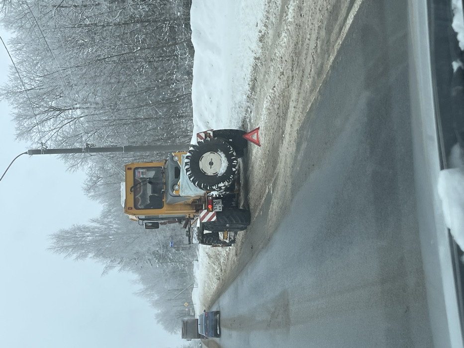 The image depicts a winter roadside scene in Belavino, Moscow Oblast, Russia. A yellow and grey utility vehicle, possibly a specialized tractor or road grader, is parked partially on the wet, dark asphalt road and partially on the snow-covered shoulder, facing away from the camera. Its rear license plate, "5745 XX 50", is visible. A red reflective warning triangle is placed on the roadside behind the vehicle. The utility vehicle features large, knobby tires, reflective striped markings on its rear fenders, and a spare tire mounted at the rear. The front section of the vehicle, including what appears to be a front-mounted attachment, is covered with a white tarp. The environment is characterized by heavy snow on the ground and covering the bare branches of dense trees lining the right side of the road, suggesting icy conditions. A tall grey streetlight pole stands to the right of the utility vehicle. In the far distance on the left, a dark blue sedan and a grey truck are visible moving away from the camera on the same road. No people are visible in the scene. The sky is overcast and appears hazy. The foreground shows the interior edge of a vehicle's windshield, indicating the photograph was taken from inside a car.