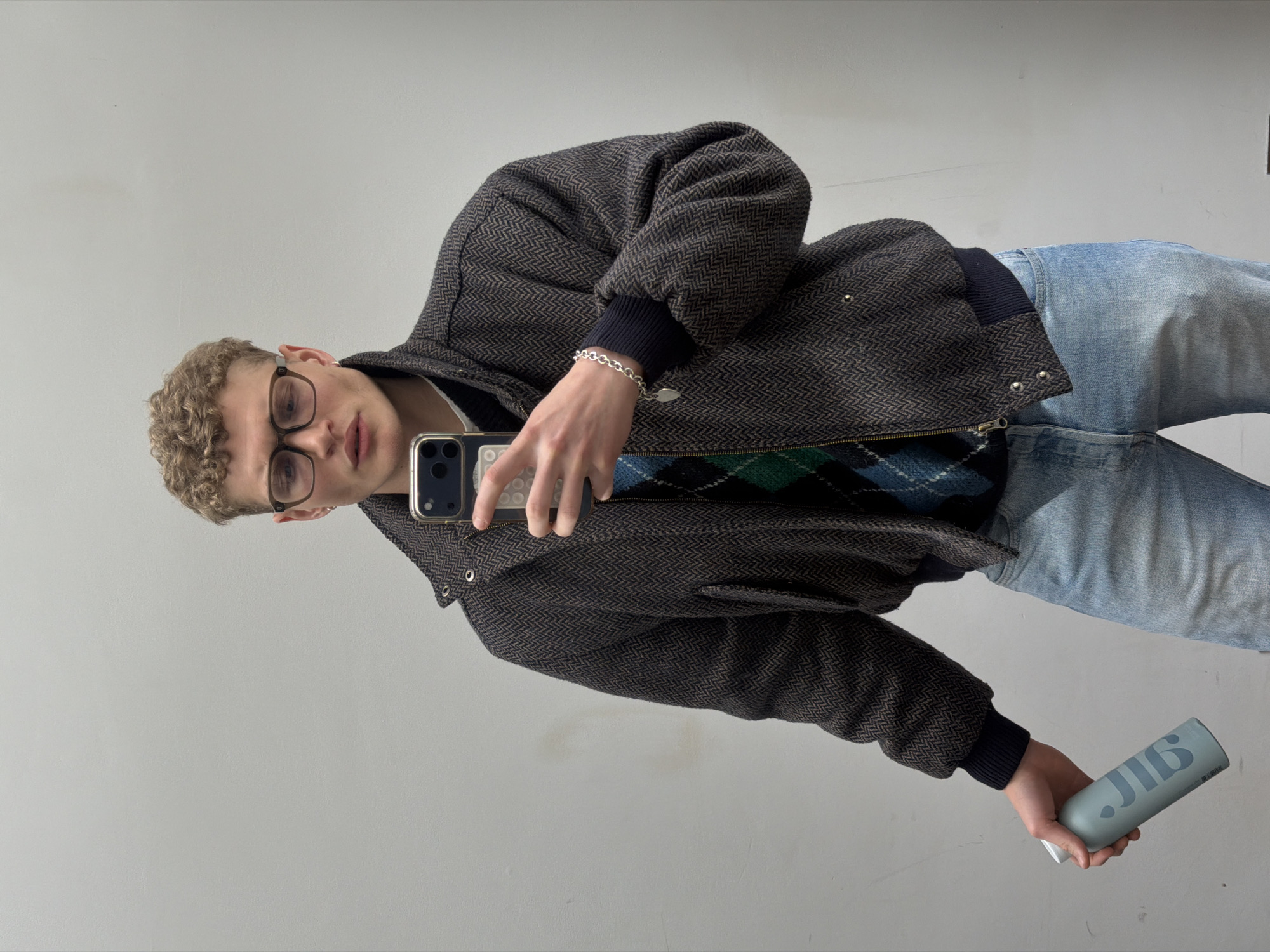A young, fair-skinned man is seen indoors, likely taking a selfie. He has curly blond hair and is wearing glasses. He is clad in a brown patterned jacket over a dark green and blue argyle sweater, and light blue jeans. He is holding a smartphone in one hand, and a light blue can with a white label in the other. The background is a plain, light grey wall. The image shows no indication of London, United Kingdom.