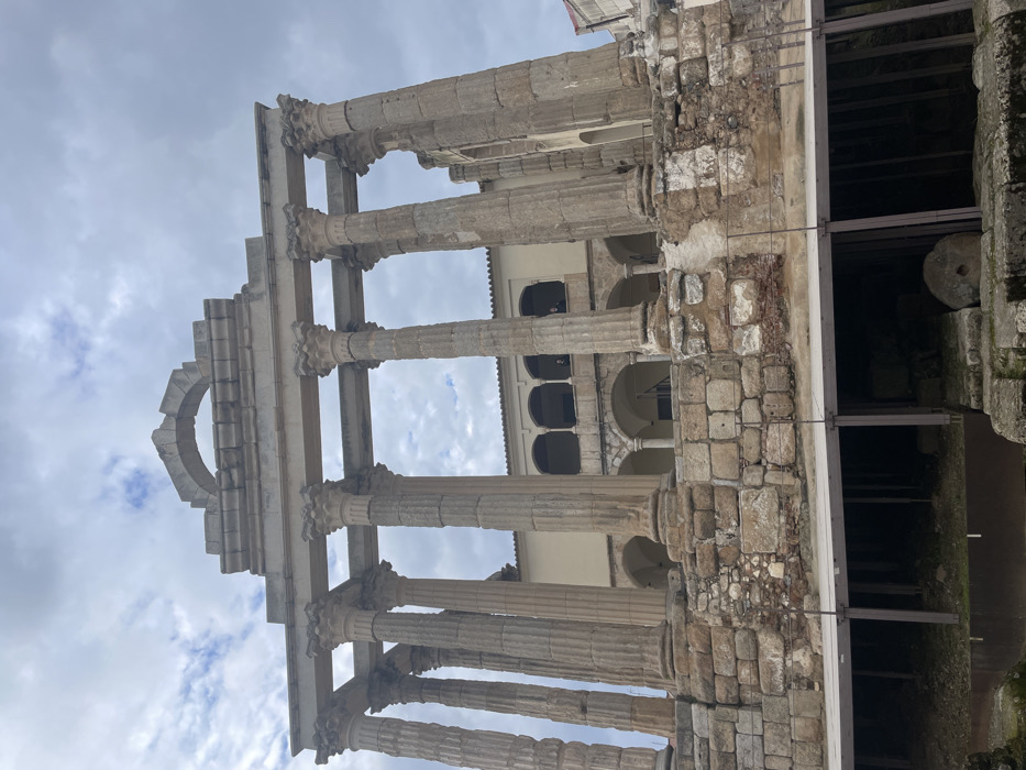 This image captures the ancient Roman ruins of a temple, identified as the Temple of Diana in Mérida, Spain. The photograph is taken from a low-angle perspective, looking up at the monumental stone structures against an overcast sky.
The primary subjects are a series of tall, weathered stone columns, likely of the Corinthian order, distinguished by their fluted shafts and intricately carved capitals. These columns support a partially preserved entablature, which includes a fragmented pediment. A prominent architectural feature is a distinct, half-arch element on the upper left side of the entablature.
To the right of the main column structure, a section of an older, robust stone wall or foundation is visible, constructed from irregularly shaped, interlocking blocks. Beyond this wall, an excavated area appears dark and shadowy, containing various stone fragments and a large, circular stone object resembling a millstone or similar ancient implement.
In the background, positioned behind the ancient temple remains, is a more modern, light-colored building. This structure features multiple arched windows and openings on its upper levels, suggesting it may be a later construction built into or adjacent to the historical site.
The sky is largely covered with light and dark grey clouds, indicating an overcast daytime scene, though some lighter areas suggest patches of less dense cloud cover. No people are visible, and no specific activity is taking place within the frame. The scene evokes a sense of historical depth and architectural endurance. There is no visible text in the image.