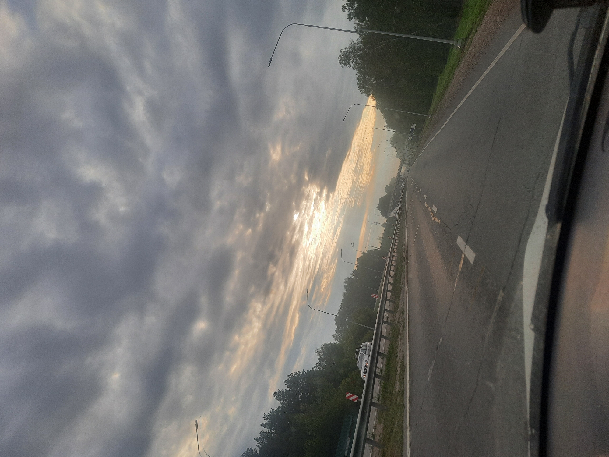 The image shows a view from inside a vehicle traveling on a highway. The sky is overcast with dramatic clouds, and the sun is partially visible through a gap, casting a warm glow. Trees line the sides of the highway, and there are streetlights and a guardrail. A white SUV is visible on the highway ahead. The perspective suggests the vehicle is moving at a moderate speed. The scene appears to be a rural or suburban highway, likely outside of a major urban center. There is no specific visual information in the image that would indicate the location is the city of Barnaul, Russia.