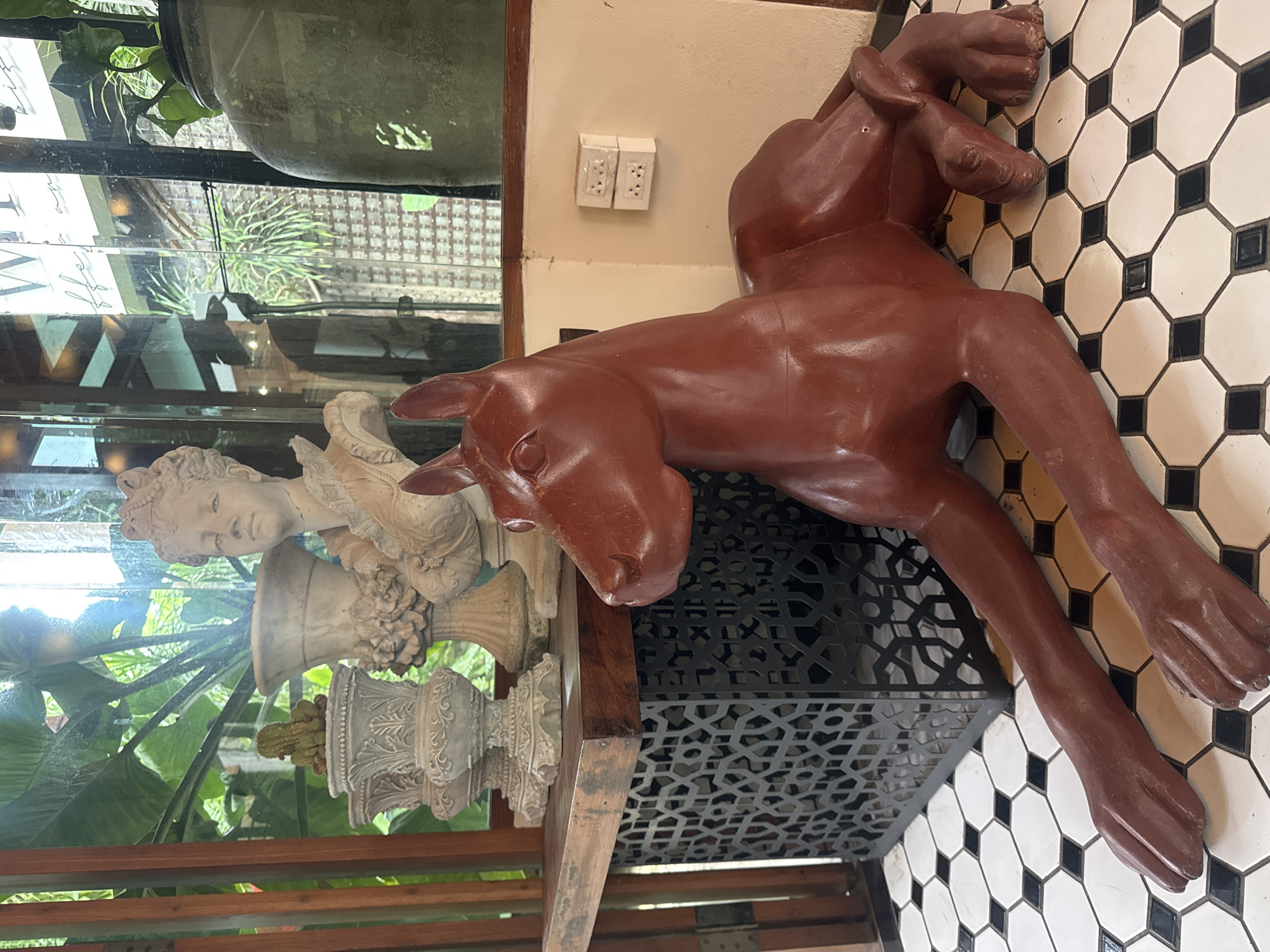 The image depicts the interior of a cafe in Suthep, Thailand, featuring a prominent, dark red, life-sized statue of a seated dog. The statue is made of a smooth, solid material and has a regal, stylized appearance, with its front legs extended forward and its head turned to the left. The dog statue is positioned on a black and white tiled floor with an octagonal pattern.
Behind the statue, a wooden table is visible, supporting several decorative items. These include a bust of a woman with an ornate headdress, a large, classic-style urn, and a smaller ornate planter containing a cactus. To the right of the dog statue and behind it, there's a wall with two stacked electrical outlets.
The background reveals large windows looking out onto lush greenery, suggesting an outdoor or garden view. The overall ambiance is one of refined decor with a touch of whimsical artistry, characteristic of a cafe setting aiming for a sophisticated yet relaxed atmosphere.