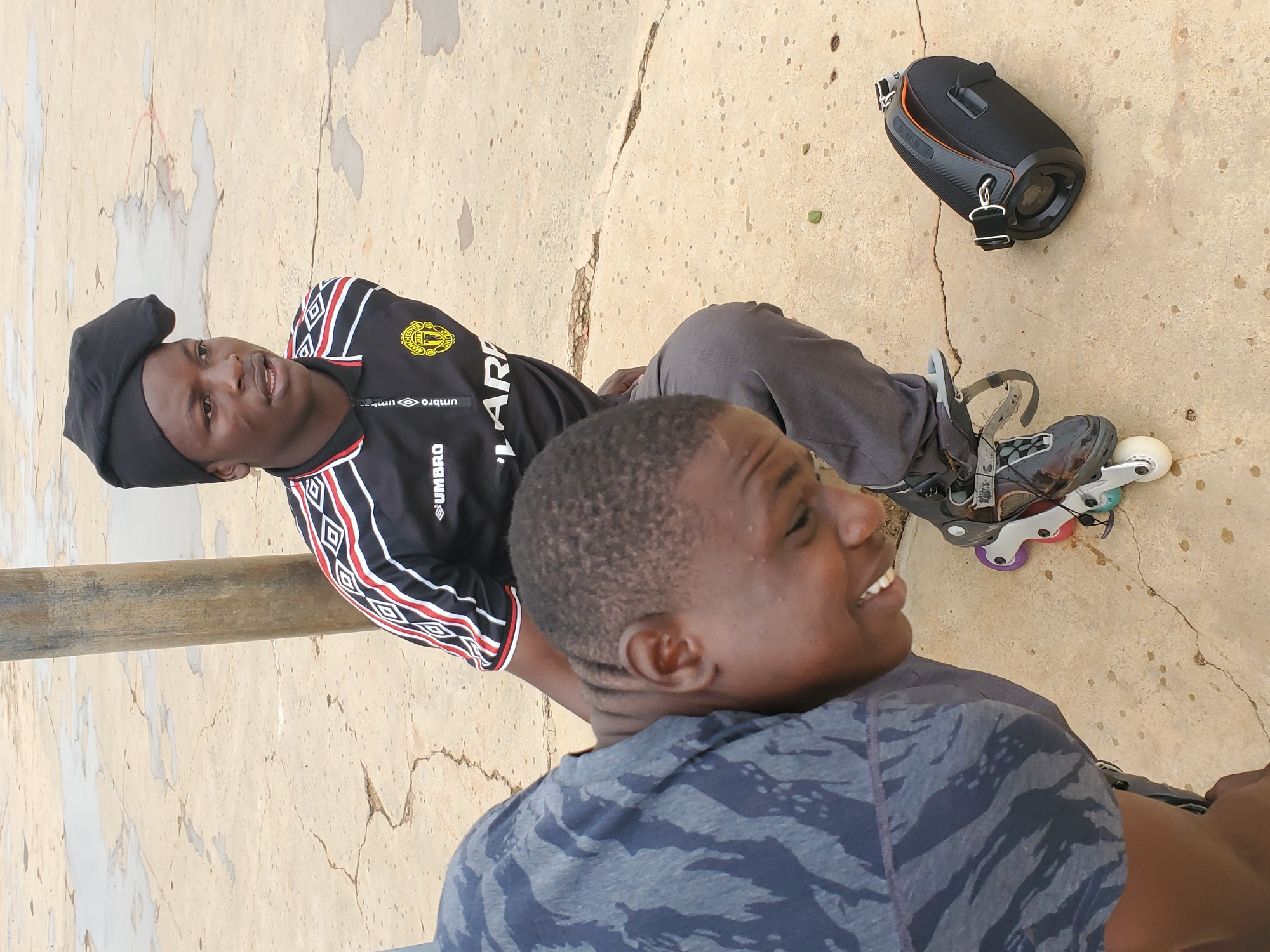 This media file shows two young men outdoors, possibly in Pankshin, Nigeria. One man, wearing a black beret and a black and red athletic shirt with white trim, is lying down with rollerblades on his feet. His shirt has the brand name "UMBRO" visible, along with a yellow crest. He is looking upwards and smiling. The second man, in the foreground, is wearing a grey camouflage t-shirt and is also smiling and looking away from the camera. He appears to be sitting or lying down, facing towards the man with the rollerblades.
In the background, a black portable speaker with orange accents rests on a textured concrete surface. The concrete appears weathered and cracked, with some scattered small, dark spots. A vertical wooden pole runs along the left side of the frame. The lighting suggests it is daytime, with soft shadows, indicating possibly late afternoon or early morning. The overall mood is relaxed and cheerful, with both individuals exhibiting smiles.