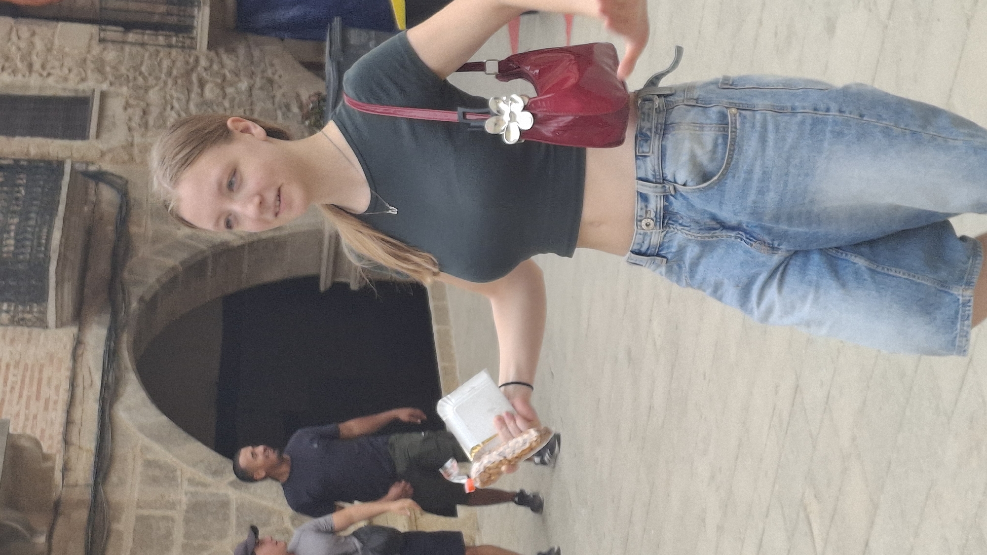 The image depicts a street scene in La Fresneda, Spain, with cobblestone paving and arched stone architecture visible in the background. In the foreground, a young woman with blonde hair pulled back in a ponytail, wearing a dark green crop top and light blue denim shorts, is walking and holding a package of food items in her right hand. She is also carrying a shiny red handbag with a white flower embellishment over her left shoulder. Behind her, two men are walking in the same direction. One man, wearing a dark t-shirt and shorts, is in the middle ground, and another man, wearing a grey t-shirt and dark shorts, is to his left and slightly further back. The overall lighting suggests it is daytime.