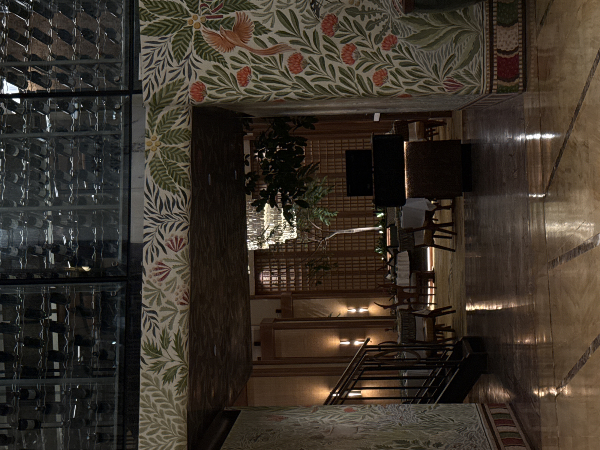 The image shows the interior of a room, likely a restaurant or hotel, with an opulent design. A large wine rack is visible on the left side, filled with bottles. Adjacent to it is a wall decorated with floral wallpaper in green, red, and beige. The interior design of the establishment features a tiered structure. Tables and chairs are arranged on different levels, suggesting a multi-level dining area. Artificial lighting fixtures illuminate the space, and a large, ornate chandelier hangs from the ceiling. The flooring is polished and reflects the light. A staircase leads to a higher level of the establishment. The overall style of the interior design is ornate and luxurious. The location is unknown, and is not necessarily in Cape Town.