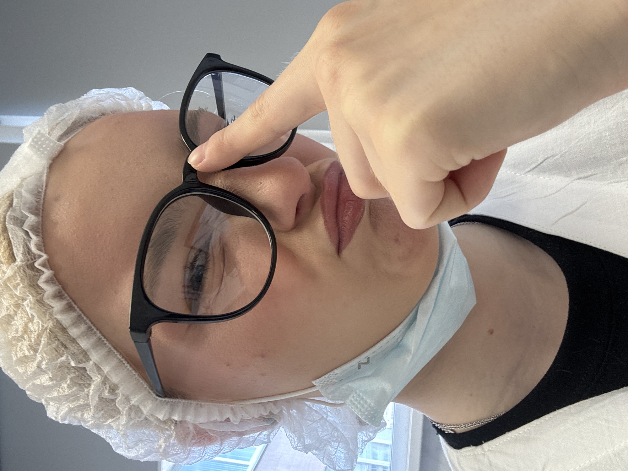 A young person with blonde hair, wearing a white hairnet, black-rimmed glasses, a blue medical mask hanging from their ears, and a white lab coat over a black shirt, adjusts their glasses with their right index finger. They are looking downwards with their mouth pursed. The background is a plain grey wall with a window on the left side showing a building. The scene appears to be indoors, possibly in a medical or laboratory setting. No specific location in the city of Unknown, Russia can be determined from the image.