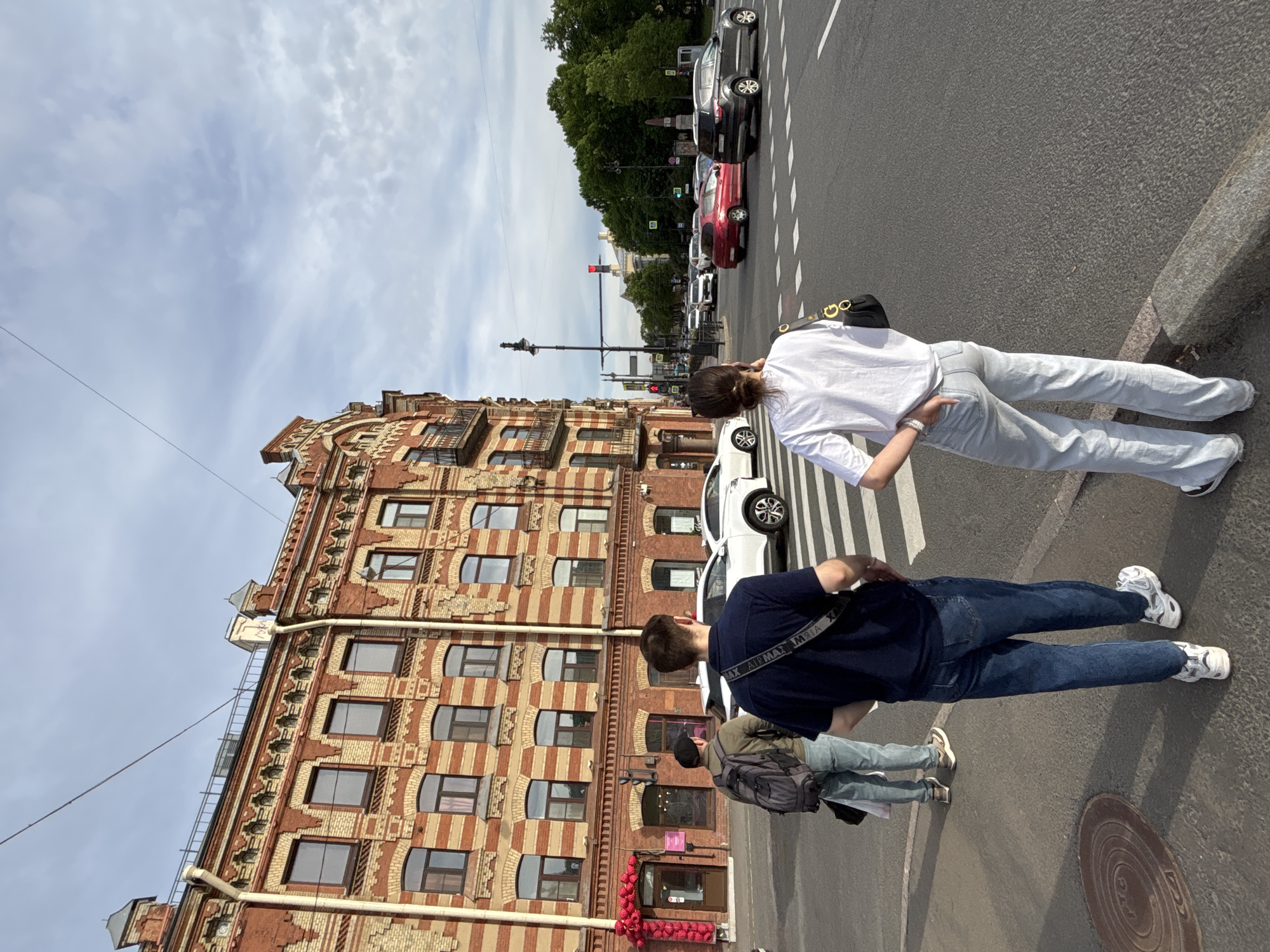 The image captures a vibrant urban street scene on a clear, bright day, rotated ninety degrees clockwise. In the foreground, two individuals are seen from behind, standing on the pavement, near a pedestrian crossing. The person closer to the camera, a woman, wears a white short-sleeved top, light-colored jeans, and white sneakers, carrying a black shoulder bag with gold-colored buckles. Behind her, a man is dressed in a dark blue short-sleeved top, dark blue jeans, and white sneakers, with a black shoulder bag displaying the text "AIK AIRMAX MIA". A third person, further back, is partially visible wearing light-colored jeans and a grey backpack.
The street is wide with a white-striped pedestrian crossing. A white car is parked near the foreground individuals, and a line of cars, including black and red vehicles, are seen further down the road, likely stopped at a red traffic light. A tall black street lamp post and the traffic light itself are prominent. Lush green trees line the street in the distance.
On the left, a striking multi-story brick building dominates the background. Its facade features alternating horizontal bands of red and lighter-colored bricks, along with numerous rectangular and arched windows. Drainpipes run along its structure. The sky above is bright blue with scattered, wispy white clouds, indicating daytime. A manhole cover is visible on the pavement in the lower right. The scene depicts a casual moment in a bustling city environment, consistent with Saint Petersburg, Russia, given the architectural style and urban setting.