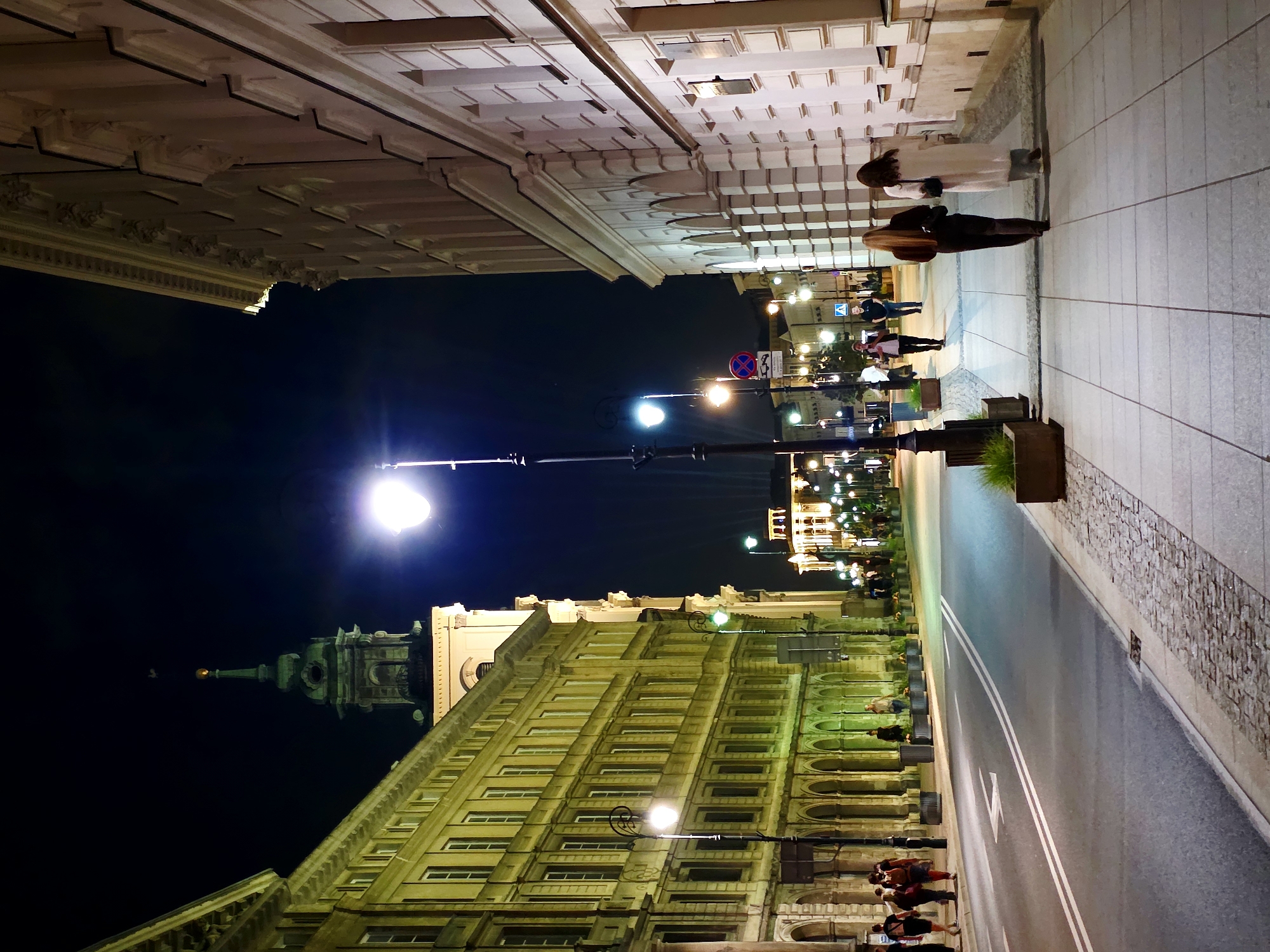 The image shows a street scene at night in Warsaw, Poland, likely on Krakowskie Przedmieście street. The view is looking down the street, with ornate 19th-century buildings lining both sides. The building on the left is illuminated with warm, yellow light, highlighting its arched ground floor and multiple windows. The building on the right is a lighter color, possibly white or cream, with a classical facade and arched windows on the ground floor.
Several streetlights illuminate the scene, casting pools of light on the wet pavement and sidewalk. The sidewalk is paved with square tiles and cobblestones. A few people are walking on the sidewalk and crossing the street. On the right side of the sidewalk, two women are walking away from the camera. The woman in front has long blonde hair and is wearing dark pants and a dark top. The woman behind her is wearing a long, light-colored coat. On the left side of the street, a group of people are walking towards the camera.
In the background, a tall clock tower with a green spire is visible, suggesting a significant landmark or church. Further down the street, more buildings are illuminated, and streetlights create a sense of depth. The sky is dark, indicating it is nighttime. The overall atmosphere is one of a bustling European city at night, with architectural beauty and a sense of movement.