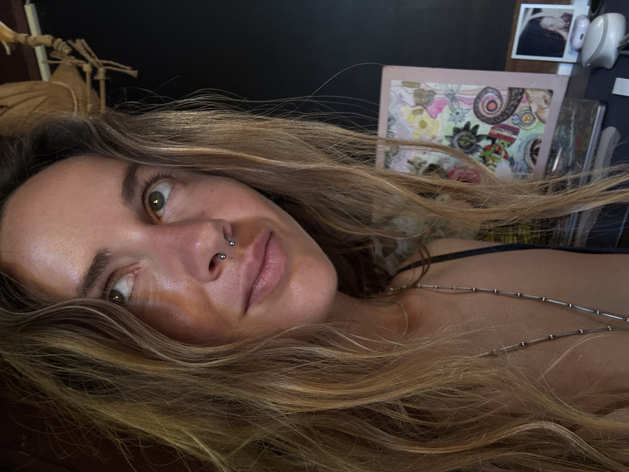 The media file is a close-up, rotated portrait of a person lying down, with their head tilted. The subject has long, blonde, wavy hair that is spread across the frame. They have a septum piercing and appear to be looking upwards and to the left of the camera. Their skin has a warm, sun-kissed glow.
In the background, several objects are visible. To the left, a portion of what appears to be a skeleton decoration, possibly a moth or butterfly, is present. Behind the subject's head, a framed artwork with colorful floral and geometric patterns is visible. Further to the right, a small framed photograph and a white electronic device, possibly earbuds, are also seen. A necklace with small silver beads is draped across the subject's collarbone. The overall setting suggests an indoor environment, with soft lighting that casts a warm tone on the subject's face. The mood conveyed is relaxed and perhaps contemplative.