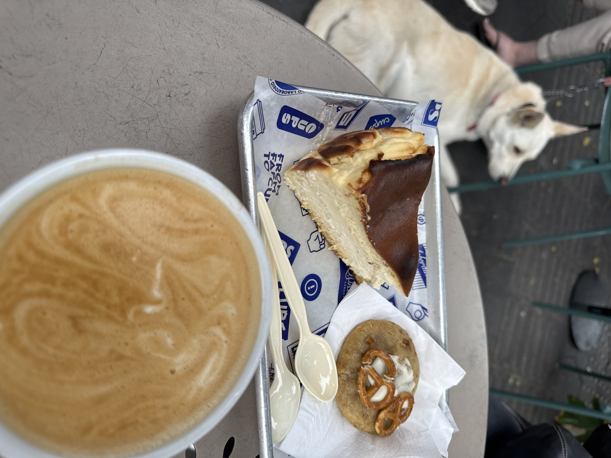 The image shows an outdoor table setting with a cup of coffee, a small tray containing a slice of Basque cheesecake and a cookie topped with pretzels and cream cheese, and two spoons. In the background, a light-colored dog is lying on the ground, possibly on a leash.
While I can identify elements typically found in Tel Aviv, such as outdoor cafes and dogs, I can't definitively say this image was taken in Tel Aviv. There are no specific landmarks or indicators in the image to confirm the location.