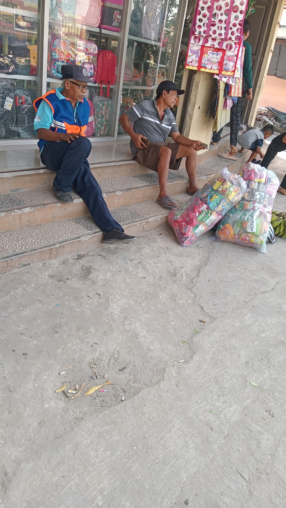 The image depicts a street scene in Kunduran, Indonesia, outside a store. Two men are seated on a set of concrete stairs. The man on the left, wearing a blue vest over a light blue shirt and dark pants, appears to be an attendant or security guard. He is wearing a dark baseball cap and glasses. He holds a small, rectangular orange object in his hands. The man on the right, wearing a grey striped t-shirt and brown shorts, is looking to his right and holds a smartphone in his right hand.
In front of the stairs, on the concrete ground, are two large plastic bags filled with various packaged food items and snacks. To the right of these bags, a bunch of bananas is visible. Further to the right, a child wearing a grey shirt is crouched down, and a person in black pants is standing.
The background shows the storefront with displays of colorful bags and other merchandise behind glass windows. The ground is made of weathered concrete, with some debris, including cigarette butts and leaves, scattered on it. The overall impression is of a bustling commercial area in Indonesia.