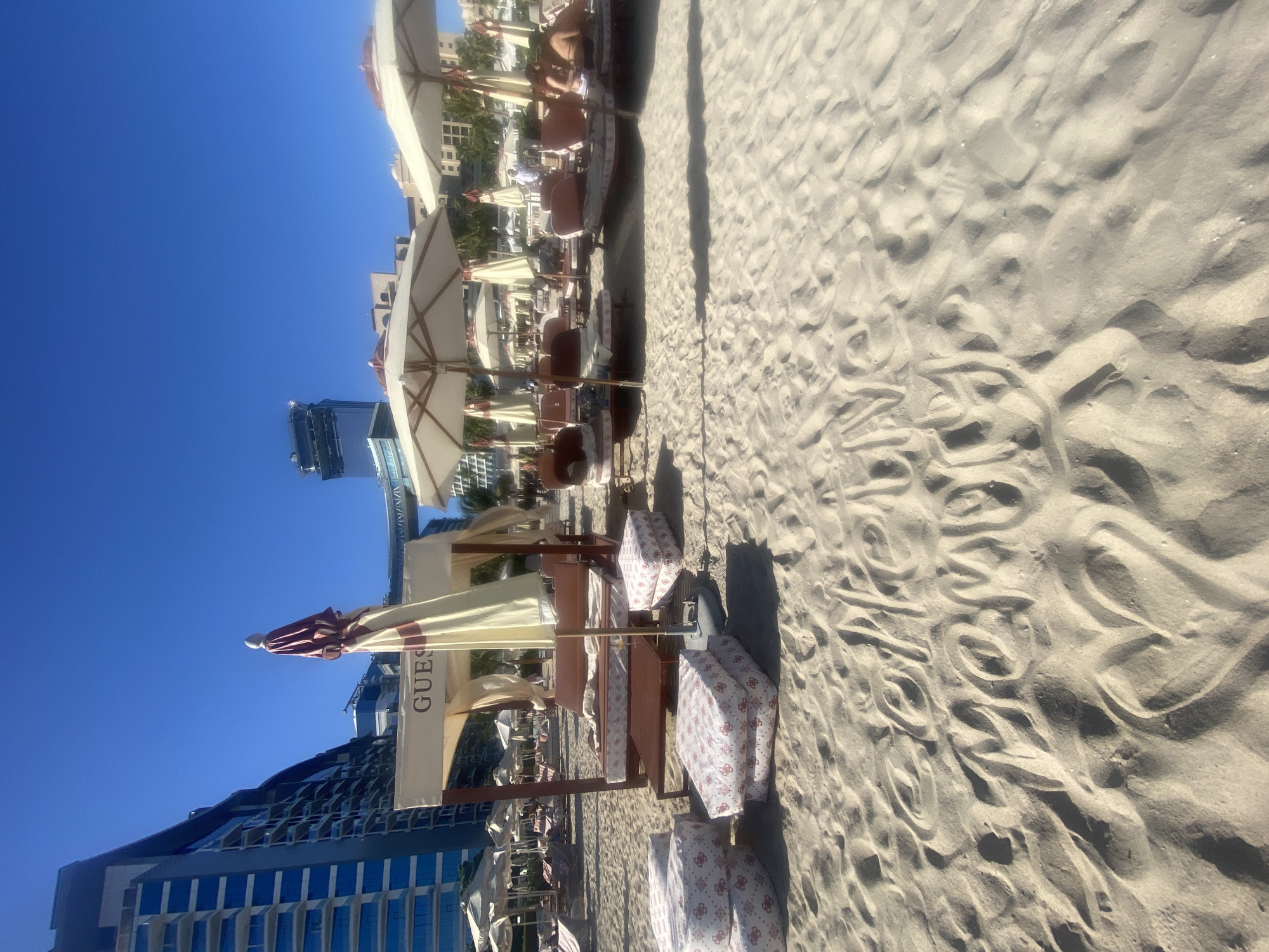 The image shows a beach scene in Dubai, United Arab Emirates. The foreground is dominated by a sandy beach with wave-like patterns. Several rows of beach loungers with white and red patterned covers are under large beige umbrellas, some of which have the word "GUESS" printed on them. In the background, modern high-rise buildings rise against a clear blue sky. Some people can be seen relaxing under the umbrellas.