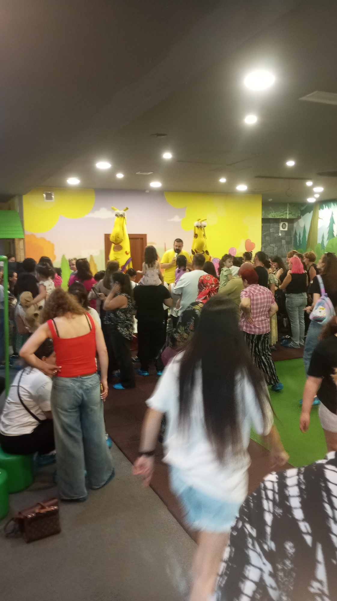 The image depicts a lively indoor event, possibly a children's party or festival, set within a brightly decorated space. The background features a whimsical mural with yellow cloud shapes against a light pink wall, and two large yellow character costumes, resembling giraffes or similar creatures, stand prominently on a raised platform. A man in a yellow t-shirt, likely an entertainer, is positioned between these characters.
A crowd of people, predominantly women and children, are gathered in the foreground and middle ground, interacting and observing the events. Some individuals are dressed in casual attire, while others wear more festive clothing. Several people are engaged in conversations, and some children appear to be playing or being held by adults.
Visible objects include a small brown handbag resting on the floor in the lower left corner of the image, and what appear to be green plastic stools. The flooring is a combination of grey and green surfaces. The overall atmosphere suggests a joyful and energetic gathering. While the specific location in Tbilisi, Georgia, is not visually identifiable from the image, the scene conveys a sense of community and celebration typical of such events.