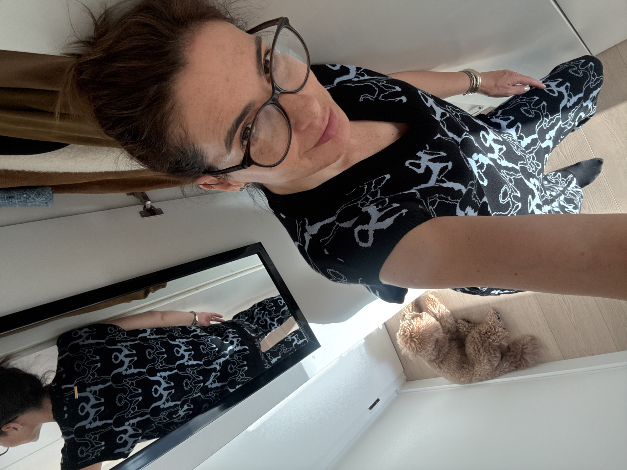 This high-angle selfie, taken in Loures, Portugal, features a woman, her mirror reflection, and a small dog in an indoor setting. The woman, appearing middle-aged with dark hair pulled back, wears large-rimmed eyeglasses and looks directly at the camera with a slight smile. She is dressed in a black top or dress and matching leggings/tights, all adorned with a distinctive light blue or grey abstract pattern. Several silver-toned bracelets are visible on her left wrist.
To the lower left, a rectangular mirror with a black frame reflects the woman's lower body and the patterned garment. The background reveals light-colored walls and a light wooden floor. A brown garment hangs partially visible on what appears to be a closet or wardrobe to the left.
On the floor in the bottom right, a small, fluffy, light brown dog (resembling a poodle) is lying on its back with its paws up, appearing relaxed. The scene is brightly lit, suggesting it is daytime. No visible text is present.