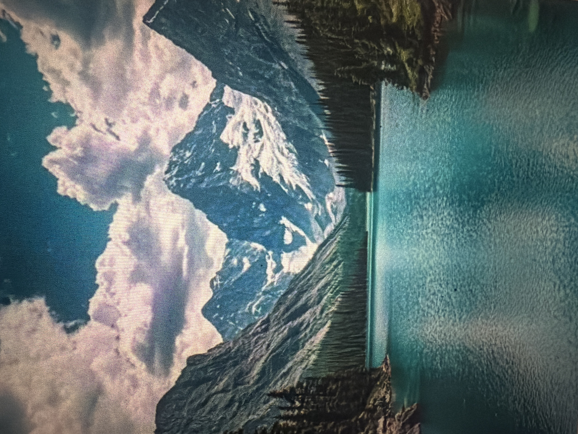 The image depicts a serene mountain lake scene. Towering, rugged mountains with snow-capped peaks dominate the background, partially obscured by dramatic, pink-tinged clouds against a deep blue sky. Dense coniferous forests line the shores of a calm, turquoise lake that reflects the surrounding landscape. The foreground is entirely filled by the tranquil water, showing gentle ripples. The image captures a pristine natural environment. The location of Baan Mae Nam, Thailand, is not present in this image.