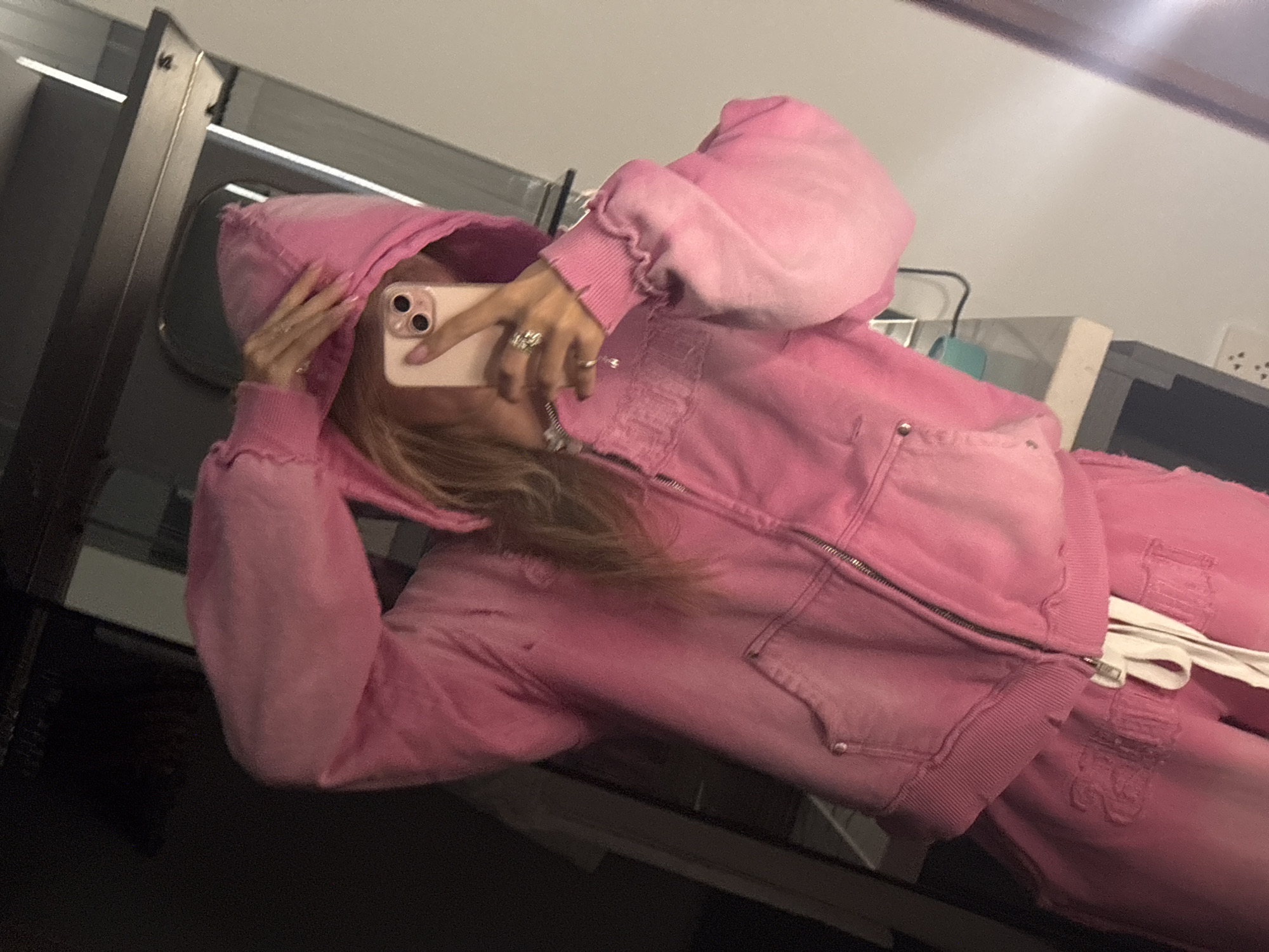 An individual, with blonde hair and visible fingernails painted pink, is reflected in a mirror. They are wearing a pink zip-up hoodie with a tie-dye effect, and matching pink sweatpants with drawstrings. The individual is holding up a pink-cased smartphone to take a selfie. The background appears to be an indoor setting, possibly a bedroom or dressing area, with a mirror, shelves, and electrical outlets visible. The overall lighting is dim.