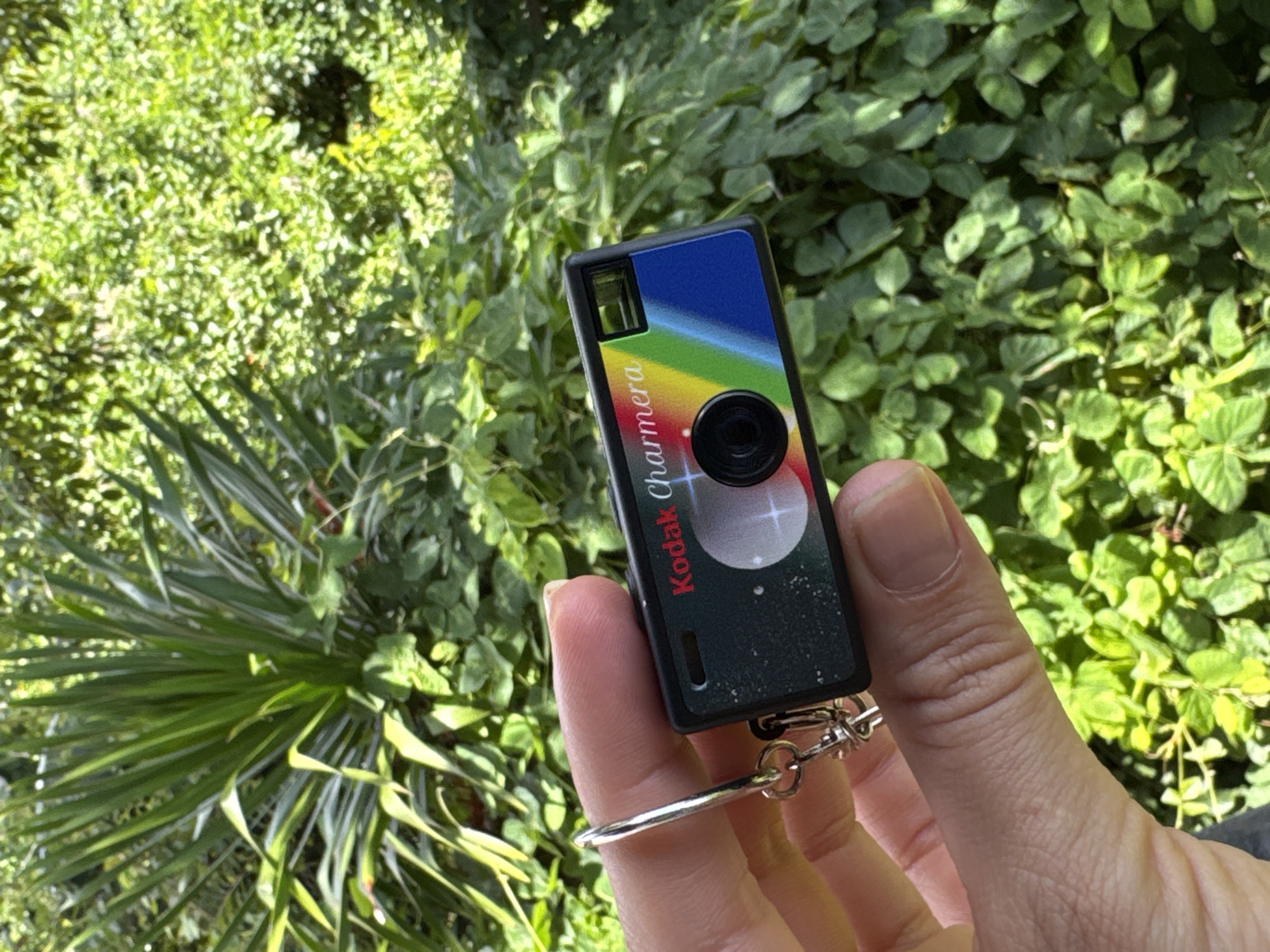 A Kodak Charmera camera keychain is held in a person's hand, against a backdrop of green foliage. The camera keychain has a blue top with a rainbow stripe and a black lens in the center. The bottom part of the keychain is dark with white speckles. The person's hand has visible wrinkles. The background vegetation includes leafy plants and spiky greenery. The location is possibly in Ungasan, Indonesia.