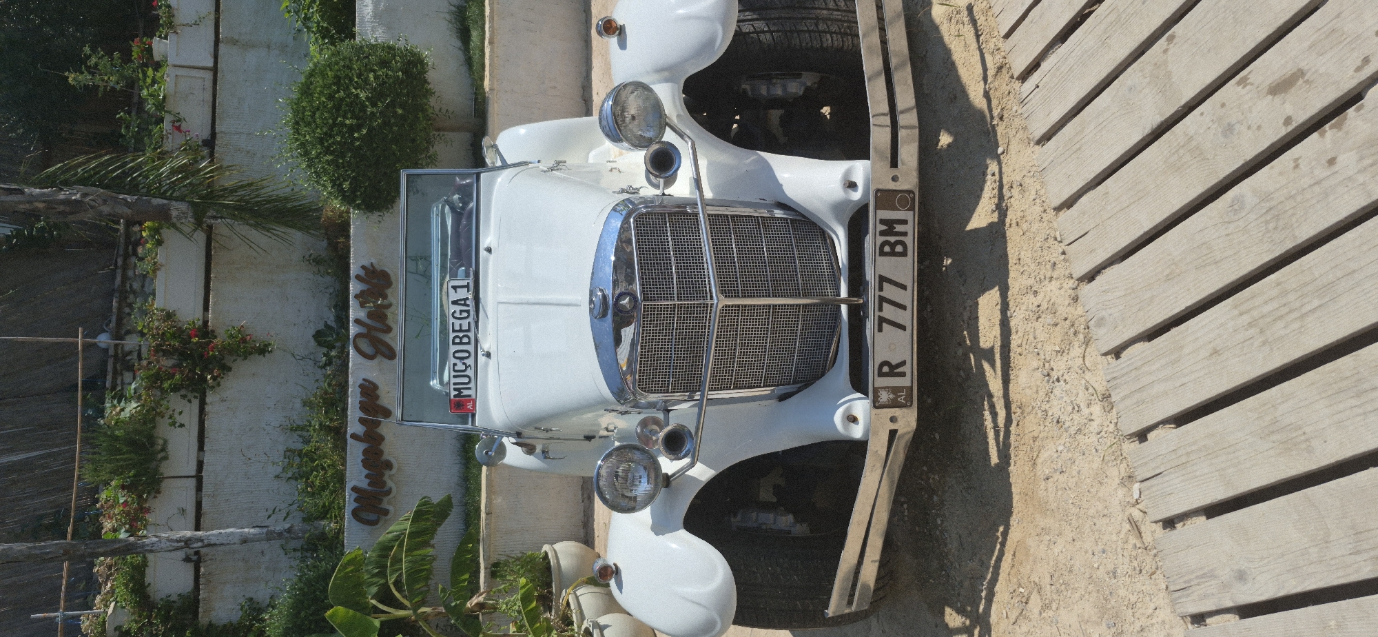The image shows a white vintage convertible car parked on a sandy ground in front of a building with a sign that reads "Mugobaga Hotel". The car has a license plate that reads "MUÇO BEGA 1" on the front windshield and "R 777 BM" on the lower front bumper. The building appears to be a hotel with potted plants and greenery surrounding it, suggesting a warm climate. The background shows a wall and some trees, and a straw-like roof structure. The overall scene suggests a hotel located in Saranda, Albania, with the vintage car possibly being part of the hotel's aesthetic or a rental service. The lighting indicates it is daytime, with sunlight casting shadows on the ground. There are no people visible in the image.