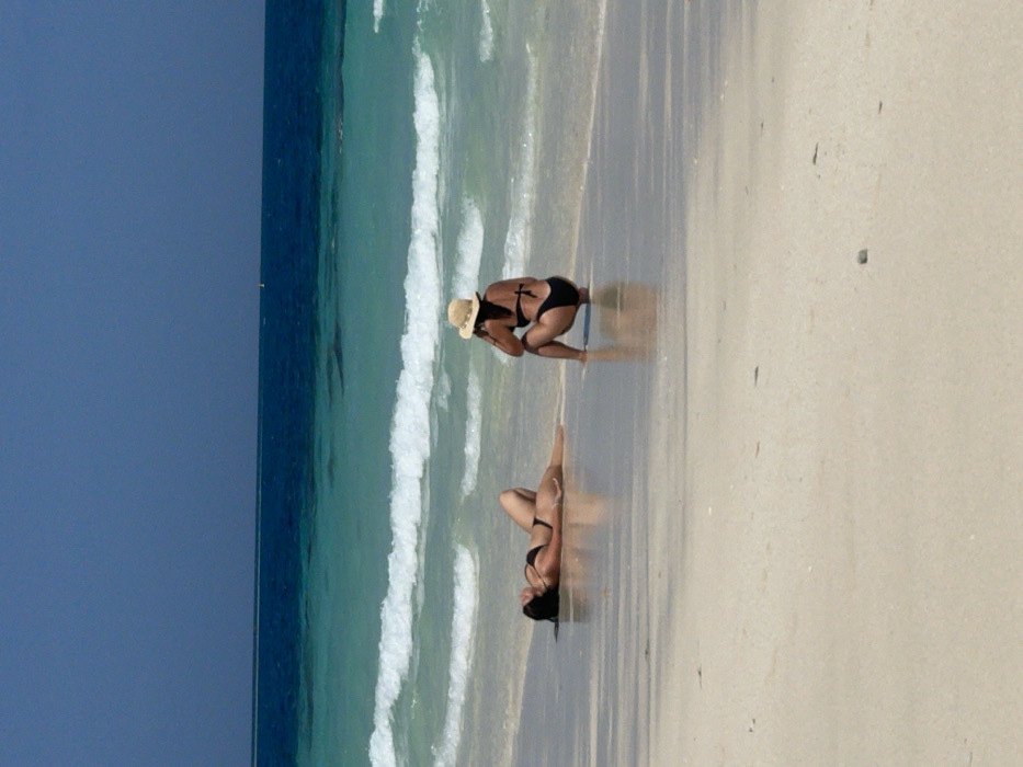 Two women in black bikinis are on a beach, with a blue sky and ocean in the background. One woman is crouching near the water's edge, wearing a straw hat. The other woman is lying on her back further up the beach. The wet sand reflects the light, creating a mirrored effect. The ocean waves are breaking in the background.