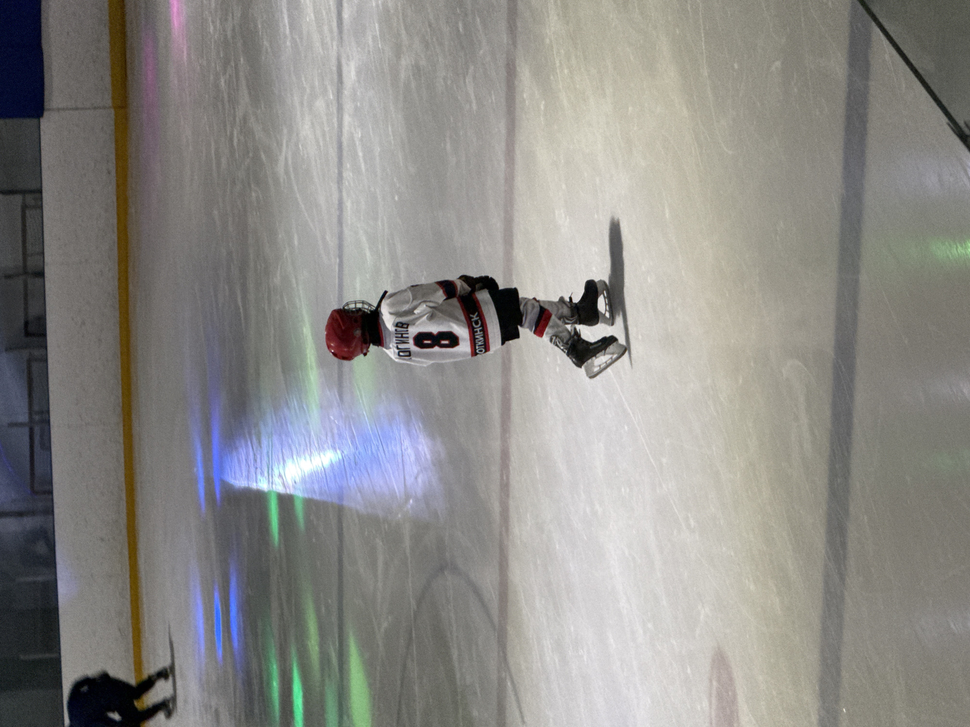 A child in a red helmet and a white hockey jersey with the number 8 and Russian text on the back skates on an ice rink. The jersey has "ОГИНОВ" across the shoulders and "ОТКИНС" across the waist. The child is wearing black hockey pants, shin guards, and skates. The ice rink has colored lights projected onto it, creating streaks of blue, green, and pink. In the background, a padded wall and lights are visible. The scene appears to be an indoor ice rink, possibly located in Votkinsk, Russia, given the text on the jersey.