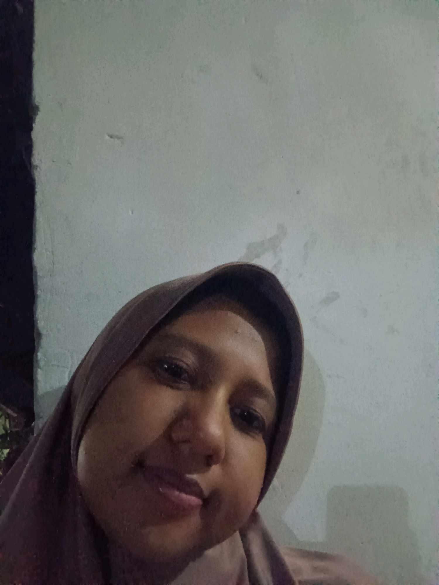 The image is a close-up, slightly distorted selfie of a woman wearing a dark purple hijab. She is looking directly at the camera with a slight smile. Her face is round, and her skin is a medium brown tone. She has dark eyes and lips. The background is a light-colored wall, appearing textured and possibly aged. To the left of the frame, a dark, out-of-focus area suggests a vertical object, possibly a doorway or a pillar, with some greenery visible behind it. The lighting appears to be artificial and somewhat dim. The image seems to have been taken indoors.