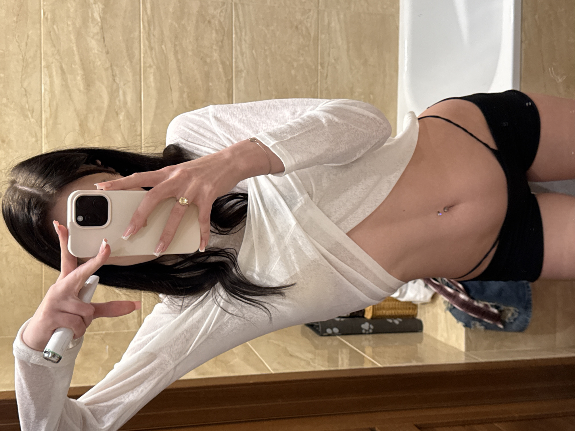 A young woman with long dark hair is taking a mirror selfie in a bathroom. She is wearing a white long-sleeved top that is tied at the waist, revealing her midriff, and black shorts. She has a belly button piercing and is wearing a ring on her right hand, which is holding up a white smartphone. Her left arm is raised, holding a white object. The bathroom has tiled walls in a sandy beige color and a white bathtub. A wooden door is visible on the left side of the frame. The location is a bathroom in Khimki, Russia.