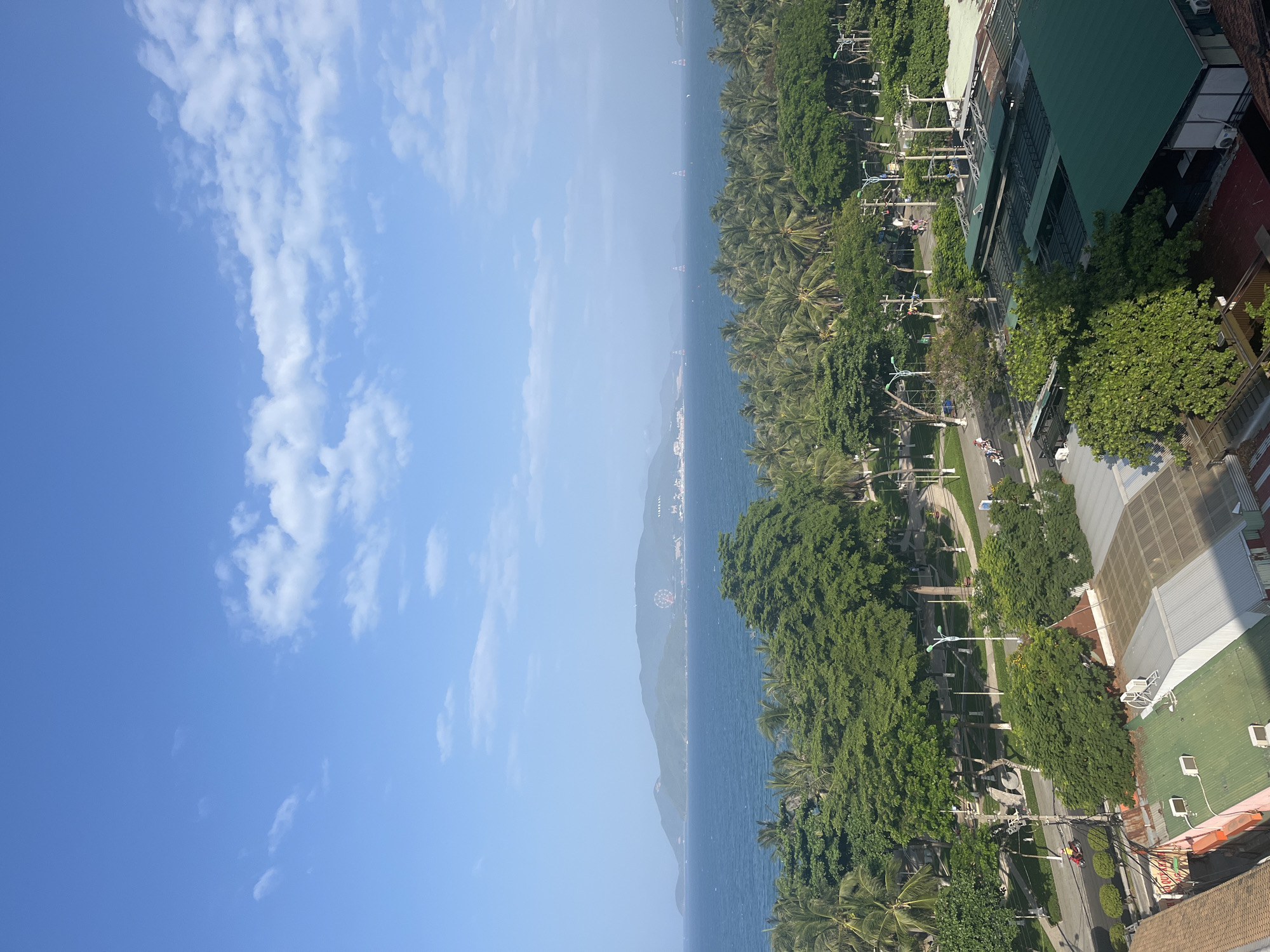 The image captures a panoramic view of a coastal cityscape in Nha Trang, Vietnam, on a clear day with scattered clouds. The foreground shows a cluster of buildings with green and brown roofs, some with visible air conditioning units. Beyond these buildings, a tree-lined street with a few pedestrians and a scooter can be observed, suggesting moderate activity. A prominent feature is a dense forest of palm trees and other lush greenery that separates the urban area from the sea. The middle ground is dominated by the calm, blue ocean, with several tall, illuminated structures, likely lighthouses or markers, spaced out in the water. In the distance, a long, mountainous island protrudes into the sea, featuring a large Ferris wheel and several buildings, indicating a resort or amusement area. The sky is a vibrant blue, providing a serene backdrop to the scene. The overall impression is one of a tropical coastal city with a blend of urban development, natural beauty, and recreational facilities.