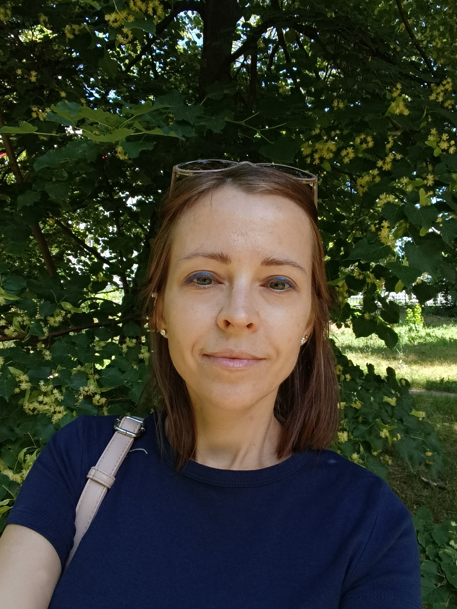 A medium shot selfie captures a Caucasian woman from the chest up, standing outdoors in front of a tree with green leaves and small yellow flowers. She has shoulder-length brown hair and is wearing a dark blue t-shirt and glasses perched on her head. A light pink strap of a bag is visible across her left shoulder. The background consists of dense foliage and dappled sunlight, suggesting a park or garden setting. Based on the provided context, the location is identified as Saratov, Russia.