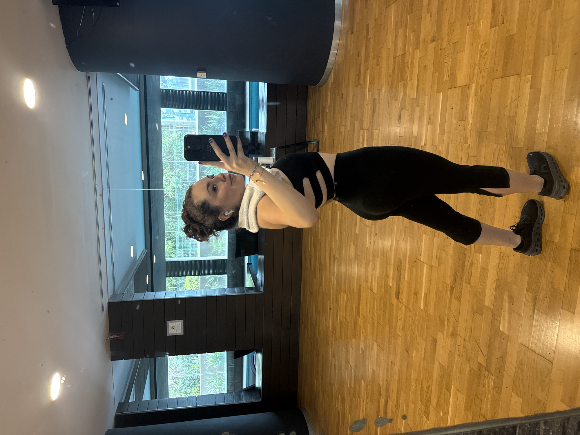A woman is standing in what appears to be a gym. She is holding a black smartphone, presumably taking a selfie in the mirror. She is wearing black workout clothes and grey athletic shoes. She has a towel draped around her neck and in-ear wireless headphones. The flooring is wooden. Outside the large windows behind her are buildings and greenery, suggesting a location within a city. Based on the decor and architectural style visible, the location is in Mexico City, Mexico.