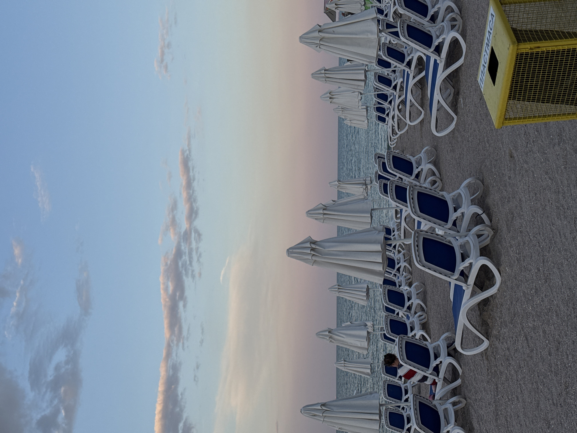 The image depicts a beach scene in Pomorie, Bulgaria, with rows of empty white beach umbrellas and blue-and-white lounge chairs on a sandy shore. The ocean stretches to the horizon under a sky with soft clouds, transitioning from blue to pink hues, suggesting either dawn or dusk. A single person, partially visible from behind, is seated on a lounge chair in the middle left of the frame, wearing a striped shirt. In the foreground, a yellow metal trash bin with a grid pattern is positioned on the sand. The overall impression is one of a quiet, possibly early morning or late evening, beach ready for visitors.