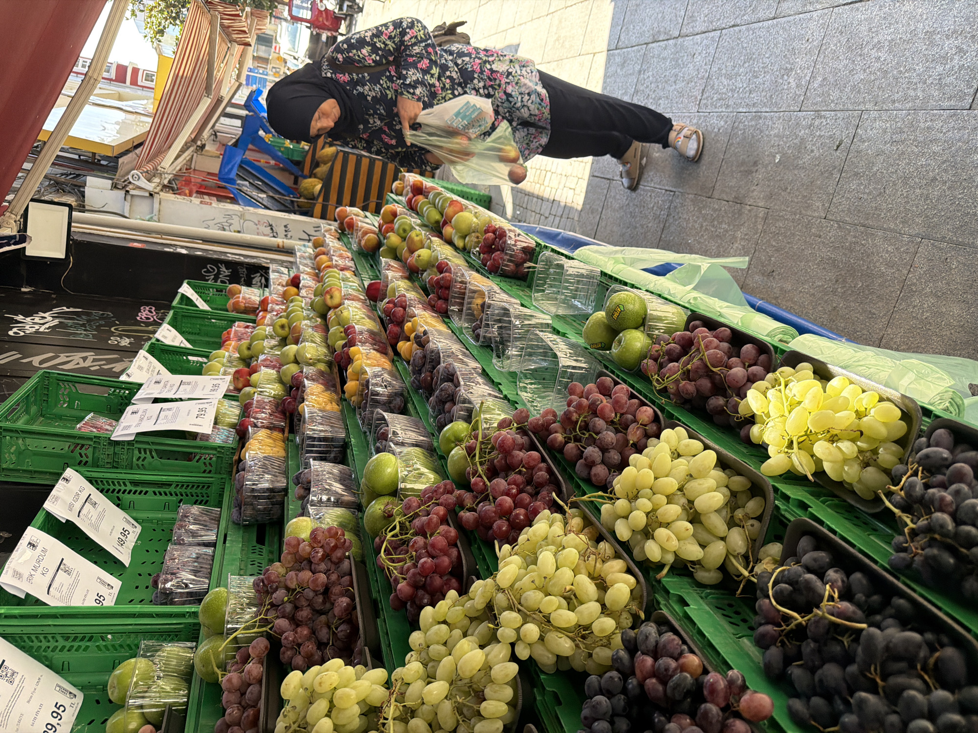This image captures a street market scene in Kadıköy, Turkey. A woman wearing a black hijab and a floral print blouse is standing on a sidewalk, looking down at a plastic bag she holds. In front of her is a display of fresh fruits, primarily grapes of various colors (green, red, and dark purple) and some apples, arranged in green plastic crates. The fruits are neatly presented in clear plastic containers and some are also loose in the crates. The background shows a building facade with graffiti and awnings, typical of a commercial street. The sidewalk is paved with granite tiles. The overall atmosphere suggests a bustling market on a sunny day.