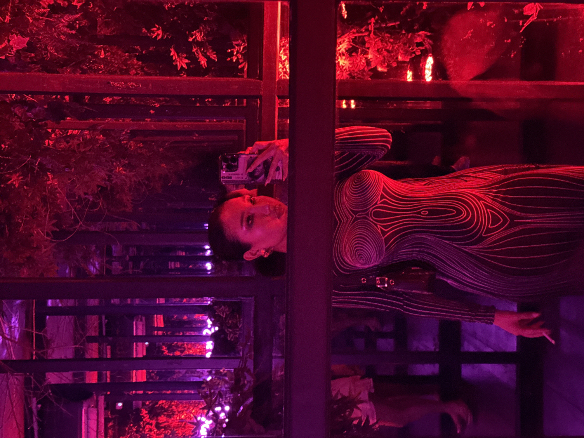 A woman is taking a mirror selfie in a dimly lit establishment in North Kuta, Indonesia. The woman is wearing a form-fitting dress with a graphic pattern resembling contours. She has her hair pulled back and is wearing large hoop earrings. In her left hand, she holds a smartphone, and in her right hand, she holds a cigarette. The background is characterized by vibrant red and purple lighting, with reflections in the mirrored surfaces. Plants and foliage are visible, adding to the atmospheric setting. The overall scene suggests a nightlife or entertainment venue.