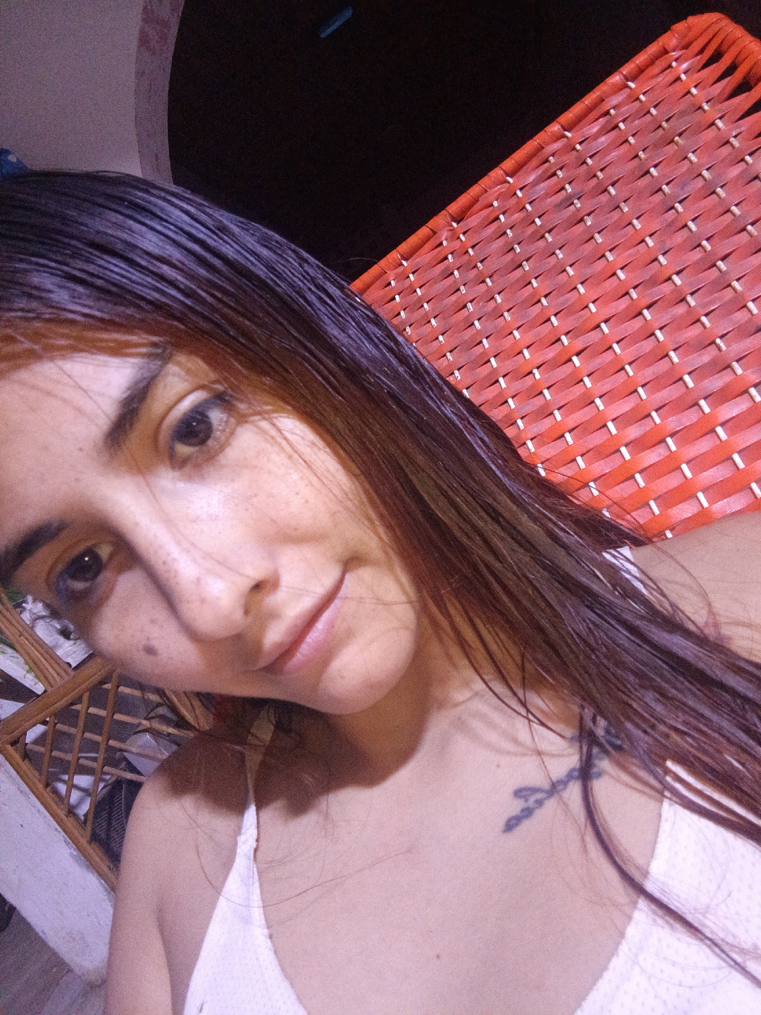 A close-up portrait of a young woman with long, straight, dark brown hair. She has light skin, freckles, and dark eyes. She is wearing a white top with thin straps. A small, dark tattoo is visible on her upper chest near her shoulder. Behind her is a woven red object, potentially furniture. A wooden structure can be seen on the left side of the frame. The lighting is somewhat dim. It is not possible to verify the specific location of Juan Griego, Venezuela from the image alone.