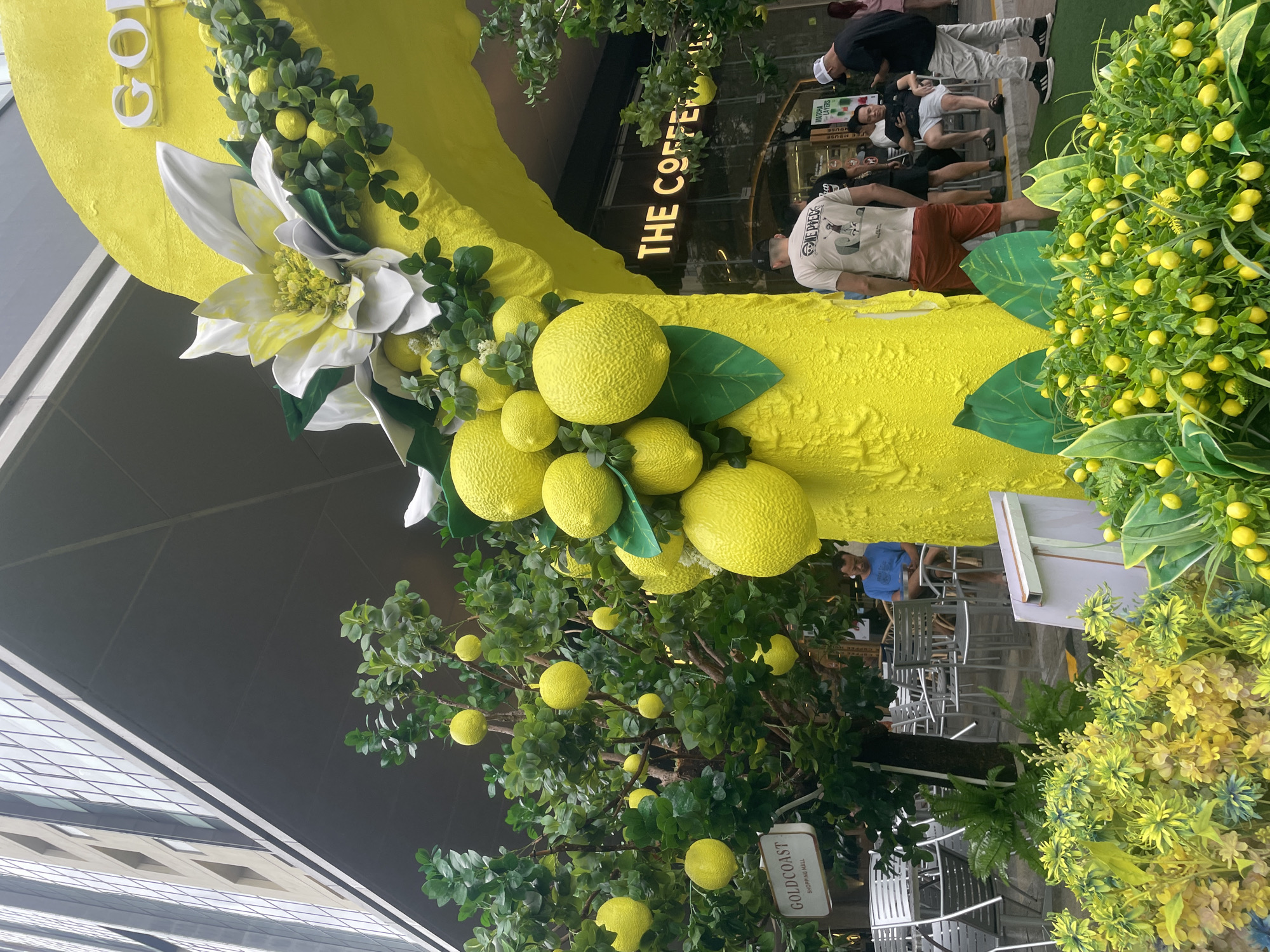 The scene shows a street-level view of a storefront, likely in Nha Trang Ward, Vietnam. In the foreground, there is a decorative archway adorned with oversized artificial lemons, leaves, and a large white flower. A sign says "GOLDCOAST SHOPPING MALL". To the right, there is a dense arrangement of artificial lemon-bearing foliage.
Behind the archway, the storefront, labeled "THE COFFEE HOUSE," has people seated inside. Outside the frame of the coffee shop, people are casually dressed and appear to be walking or seated at tables.
The surrounding environment includes a modern building with a reflective surface visible on the left side of the frame. The ground is paved, and additional seating is arranged nearby.