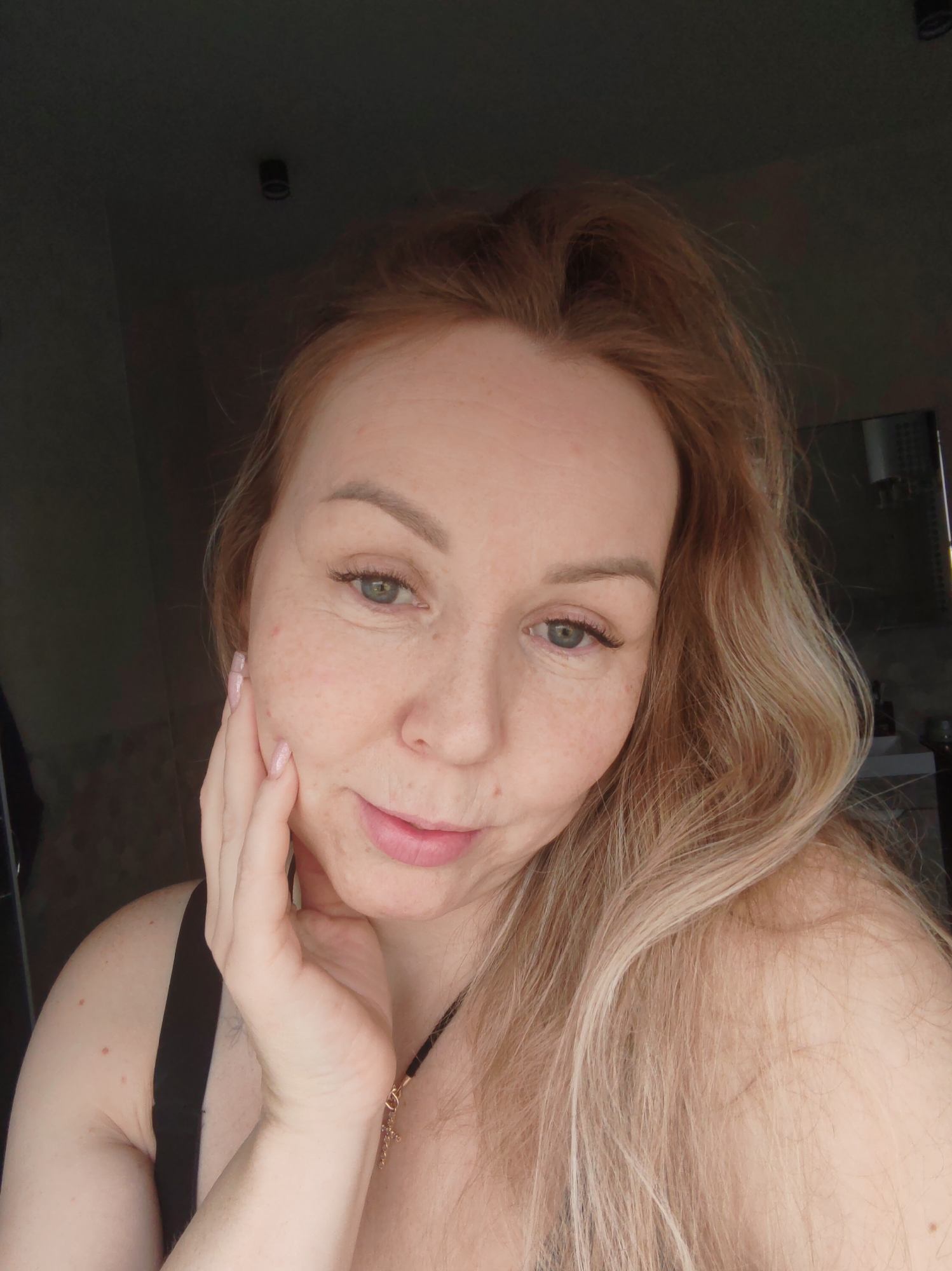 This is a close-up, slightly angled selfie of a woman, presumably taken indoors in Chaikovsky, Russia. The woman, who appears to be in her late 20s to early 40s, has fair skin with visible freckles across her cheeks and nose. Her eyes are a light blue-green, and she has naturally colored, full lips with a gentle, faint smile. Her hair is long, wavy, and multi-toned, featuring reddish-blonde roots that transition into lighter blonde strands, flowing over her right shoulder.
She is posing with her left hand resting softly against her cheek and jawline. Her fingernails are manicured with a light pink or glittery polish. She wears a black strap garment, possibly a tank top or bra, and a dark cord necklace with a small gold-colored cross pendant.
The background is dimly lit and out of focus. On the left, a dark wall is visible, while on the right, there's a reflection, possibly from a mirror or a cabinet, and a hint of a white shelf. A dark, cylindrical light fixture is dimly visible on a dark ceiling in the upper portion of the frame. The lighting suggests a natural, soft illumination coming from off-camera, possibly a window, creating a somewhat intimate atmosphere. There is no visible text in the image.