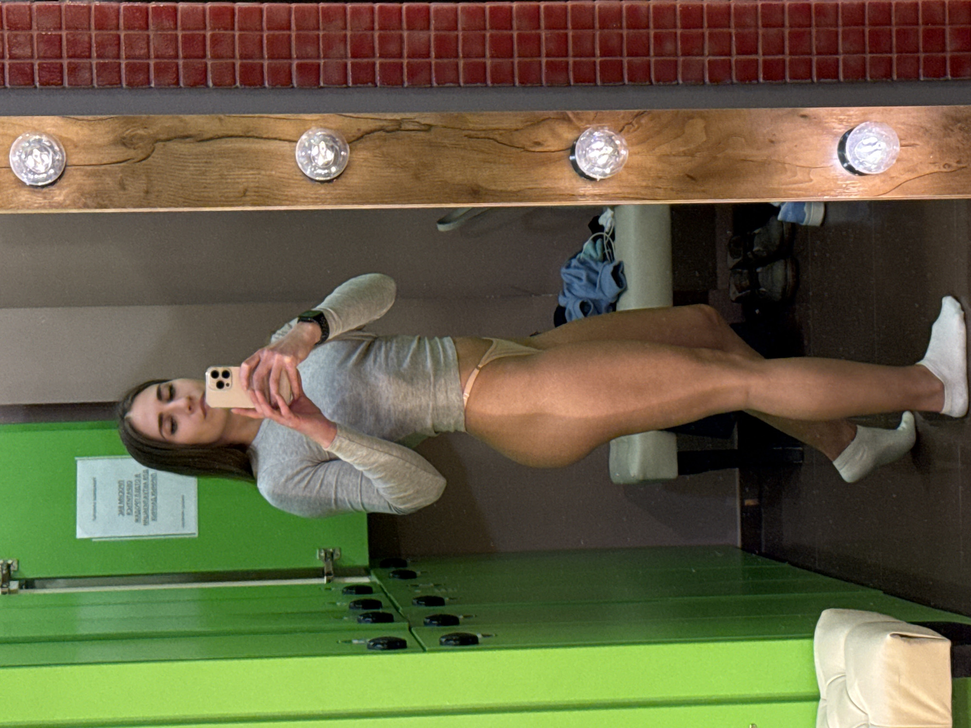 A full-length mirror selfie shows a young woman in what appears to be a locker room or changing area, reflected in a mirror. The image is rotated 90 degrees clockwise.
The woman, with long dark hair, stands sideways, holding a light-colored smartphone with both hands to take the picture. She is wearing a light grey long-sleeved top, light-colored (possibly beige or nude) underwear, and white socks. A smartwatch is visible on her left wrist. Her legs are tanned, with a distinct tan line visible near her hips where her underwear meets her skin.
In the reflection, above the woman, red square tiles are visible. Below these tiles is a wooden panel with three circular, decorative light fixtures that cast bright reflections. Below the wooden panel, a dark grey wall is visible. To the right of the woman's reflected body, some blue clothing is folded, and shoes are visible on a dark floor. A white cylindrical object, possibly a bin or fixture, is also seen.
In the foreground of the reflection, on the left side, are bright green lockers with black handles/locks. A white rectangular sign with text is affixed to one of the green lockers, but the text is illegible due to glare and resolution. The floor in the reflected locker room is dark.